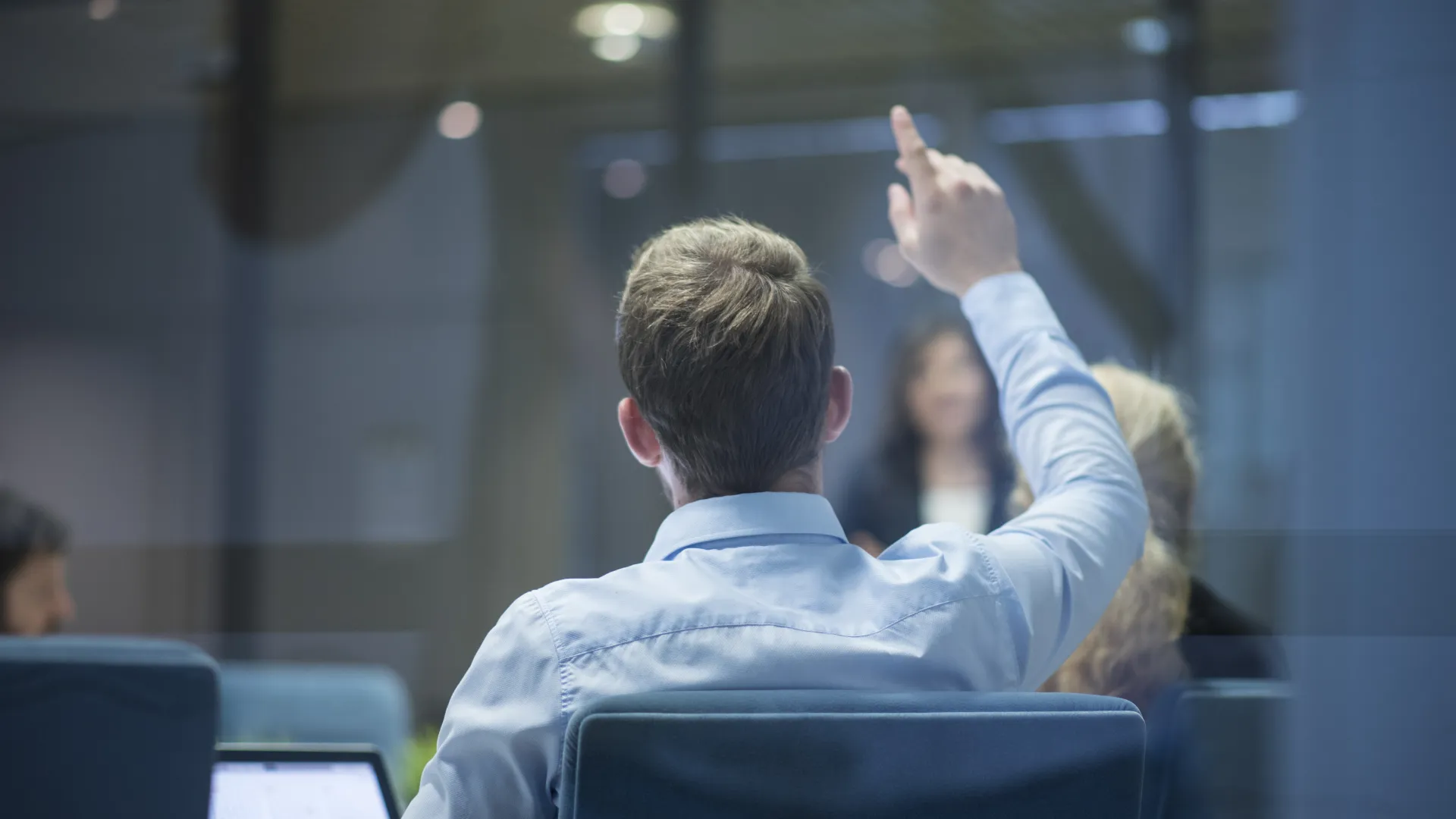 Man in office raises hand during meeting, colleagues and laptop visible in modern conference room.
