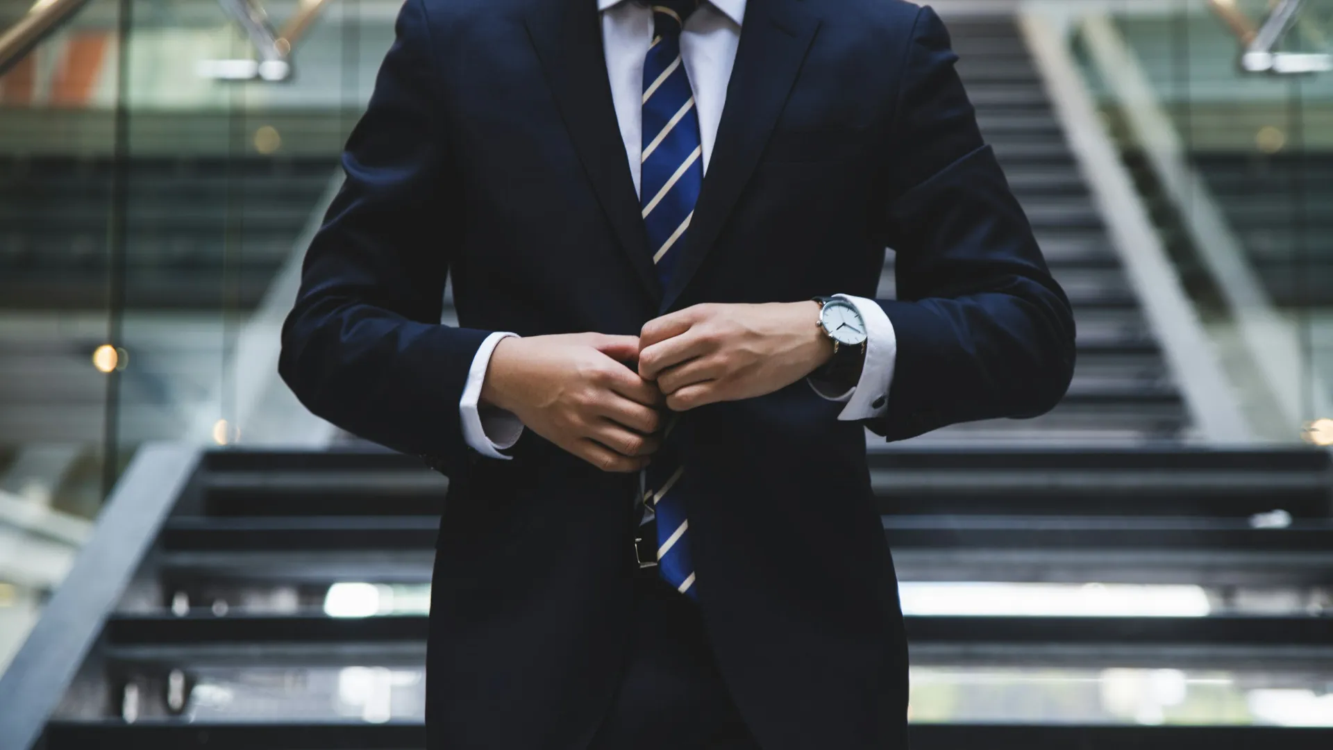 Businessman buttoning suit jacket standing in front of modern staircase wearing a tie and watch
