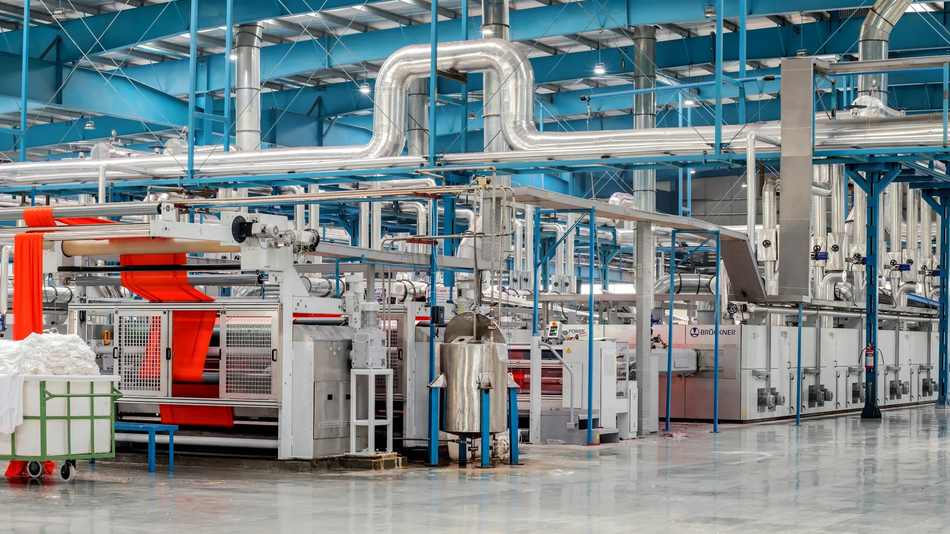 Modern textile factory interior with large automated machines, fabric rolls, and industrial piping under blue ceiling.