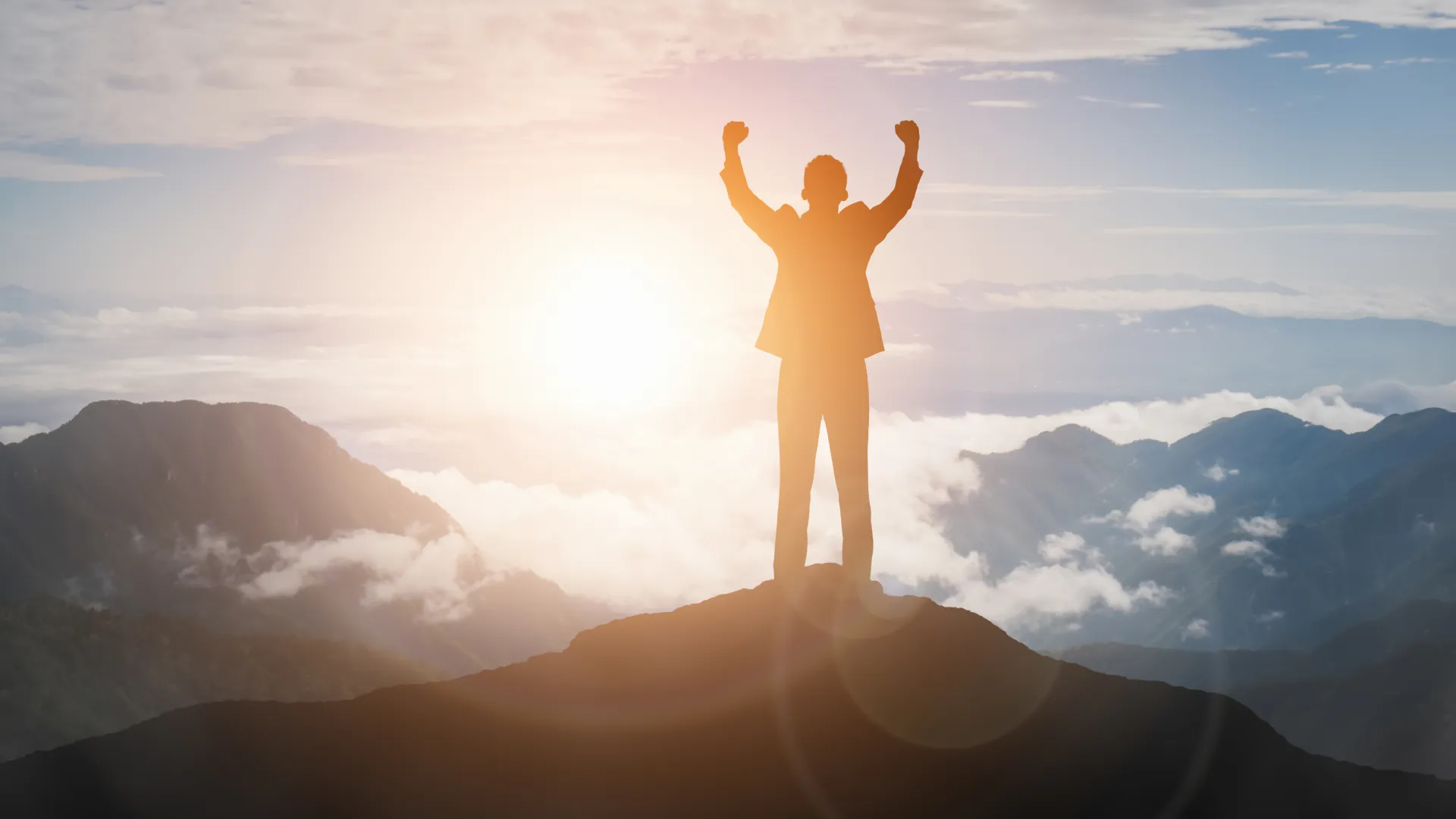 Silhouette of a person celebrating victory atop a mountain peak during sunrise with clouds and mountain ranges.