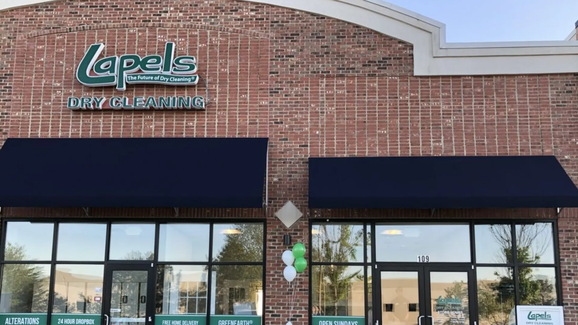 Exterior view of Lapels Dry Cleaning store with brick facade, black awnings, and promotional window signs.