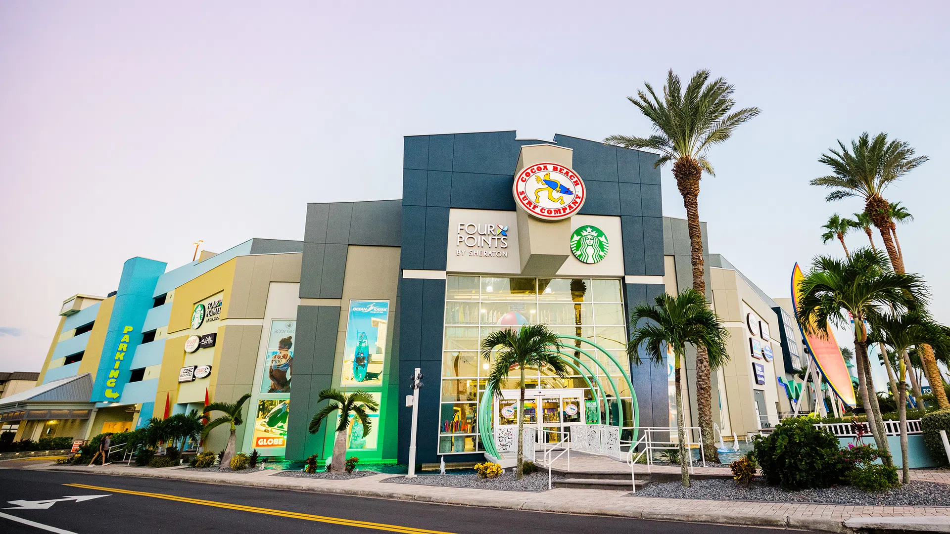 Modern shopping plaza with Four Points by Sheraton, Starbucks, palm trees, and parking garage under clear sky.
