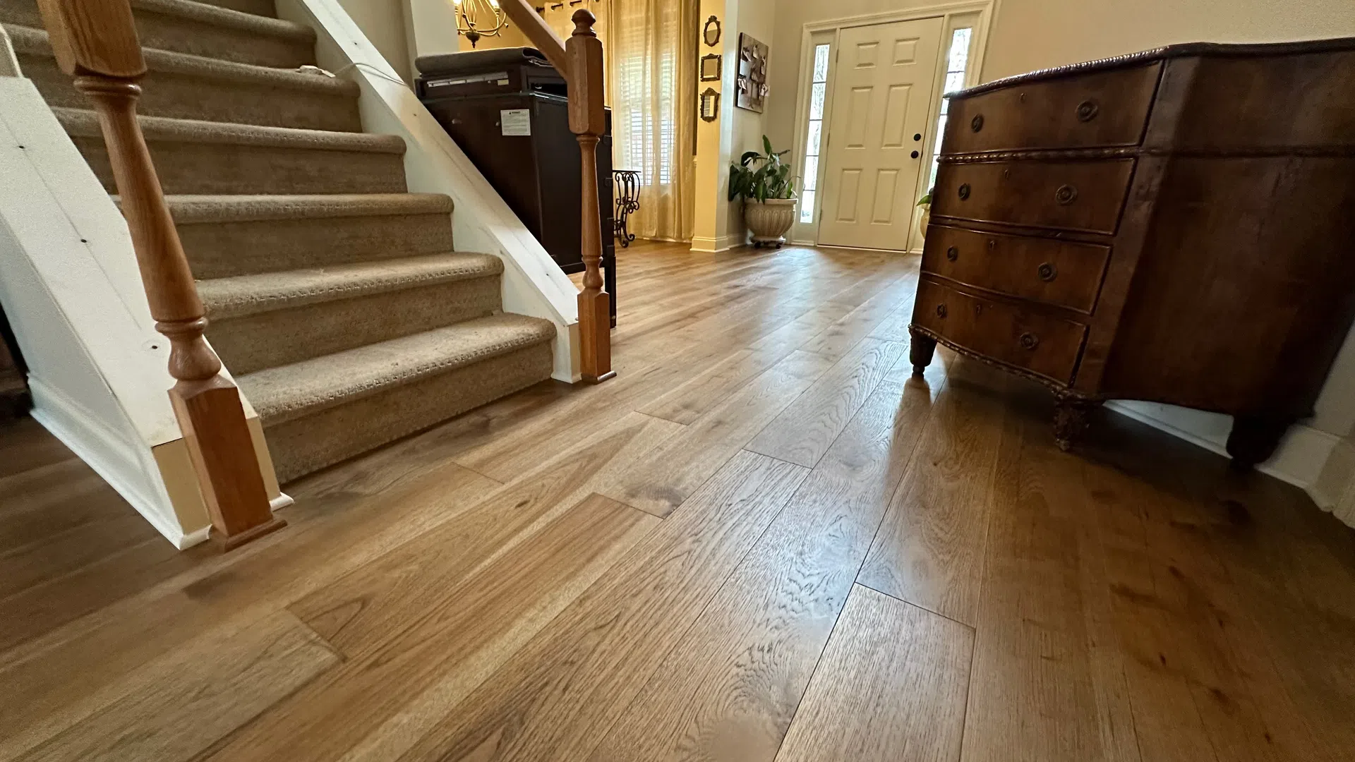 Wide-angle view of a home entryway with wooden flooring, carpeted stairs, and vintage dresser under warm lighting.