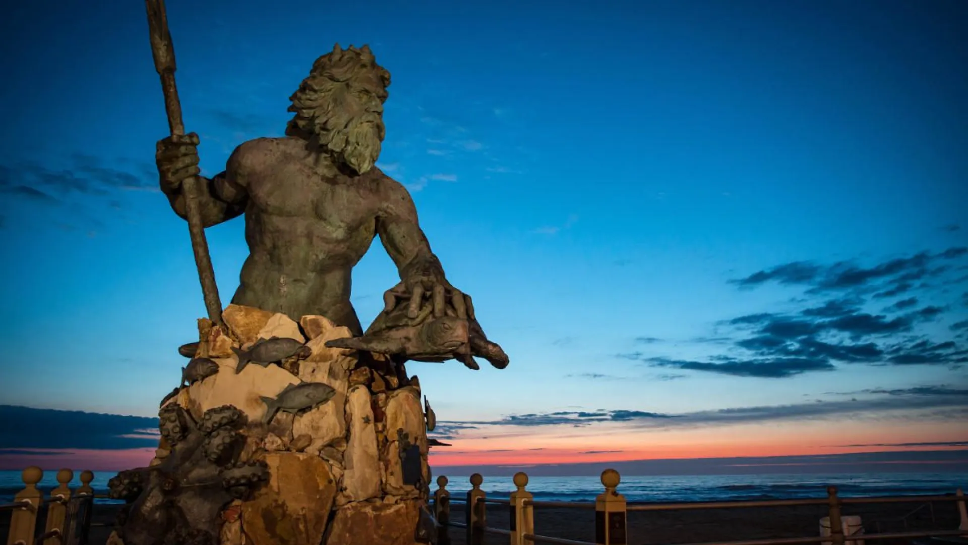 Statue of Poseidon holding a trident and sea creatures at sunset by the oceanfront.