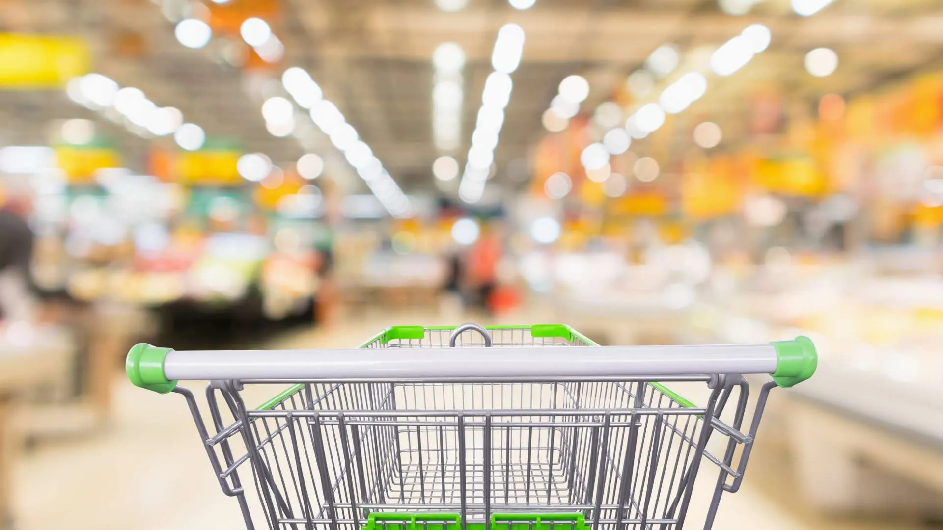 Empty shopping cart with green handles in a brightly lit supermarket aisle with blurred background.