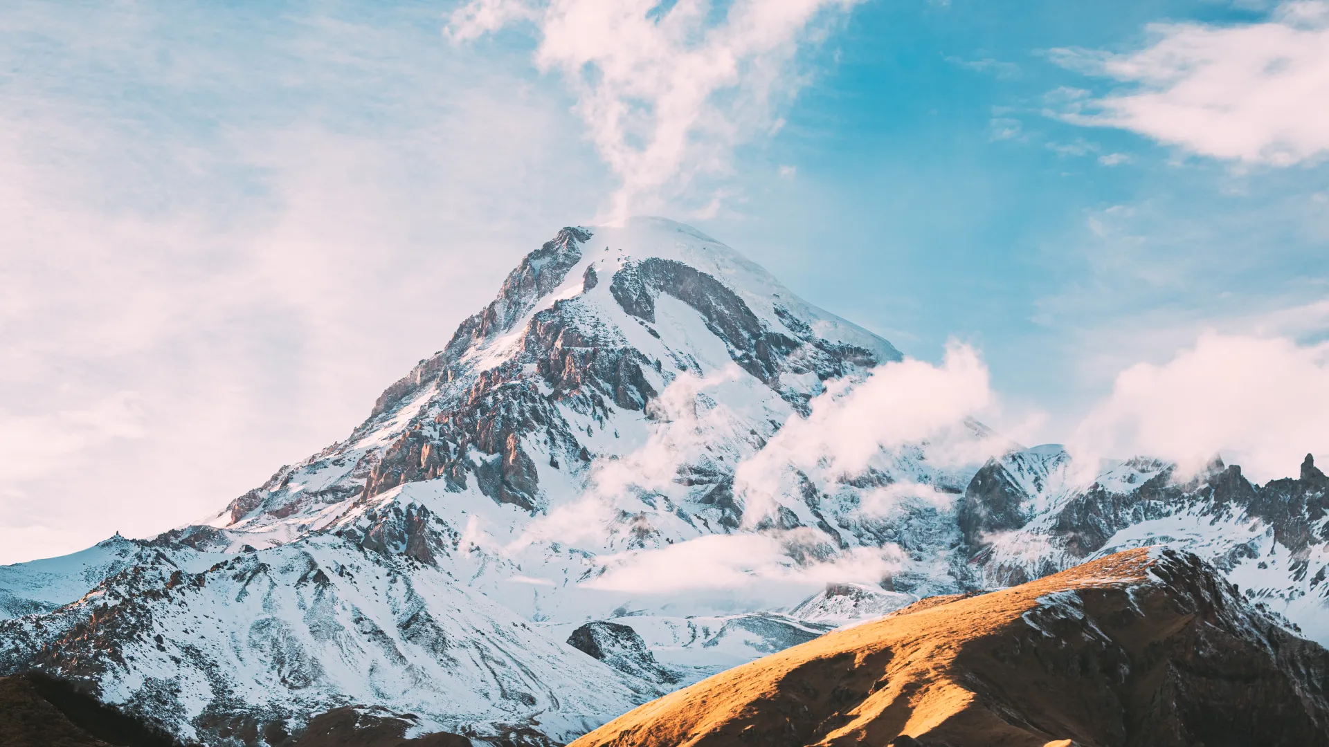 Snow-covered mountain peak with clouds drifting overhead and sunlit rocky hills in the foreground under a blue sky
