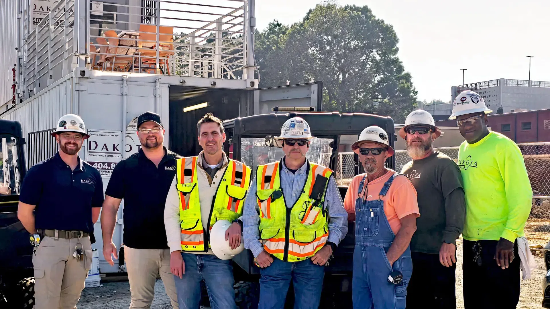 Seven construction workers wearing safety gear pose at a job site with equipment and containers in the background