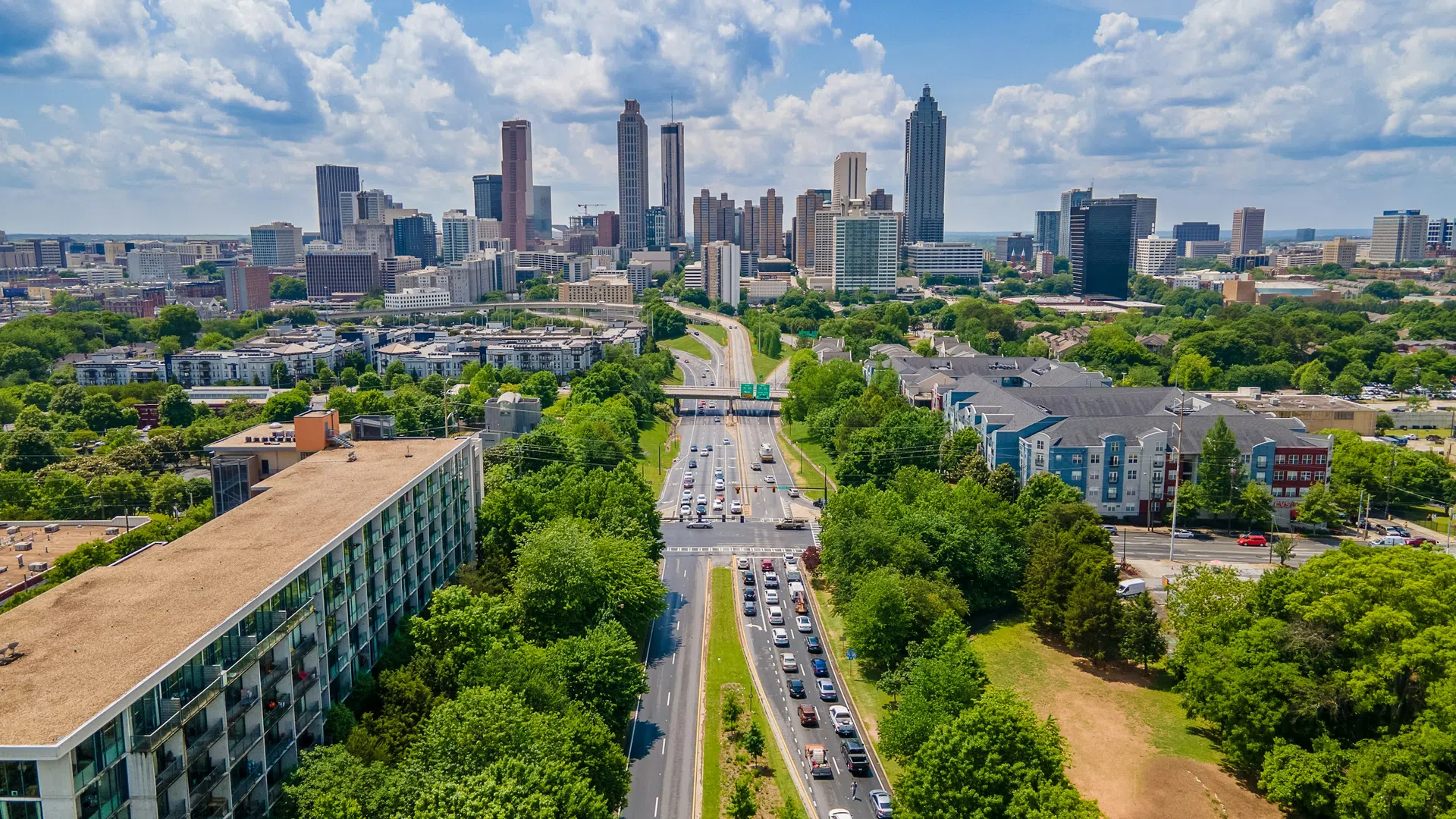 Aerial view of a highway leading into a city skyline with tall buildings under a partly cloudy blue sky.