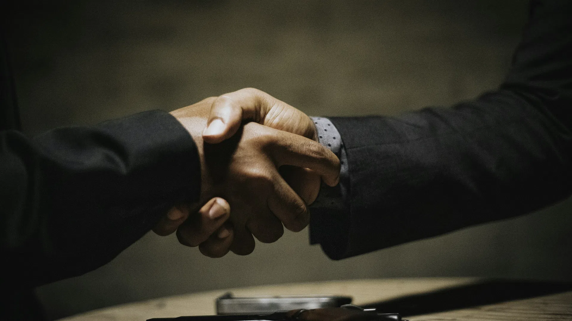 Two people shaking hands over a table with a handgun placed on it, symbolizing a tense or secretive agreement.