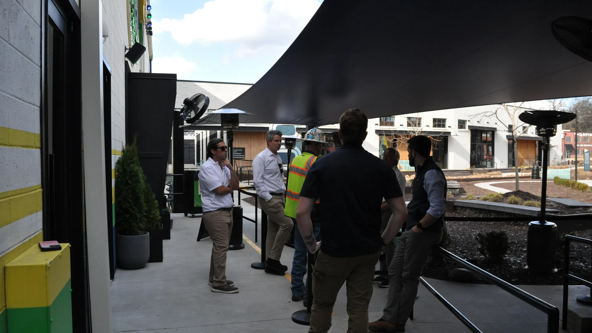 Group of people, including one in a safety vest, talking under a large black canopy outside modern buildings on a sunny day