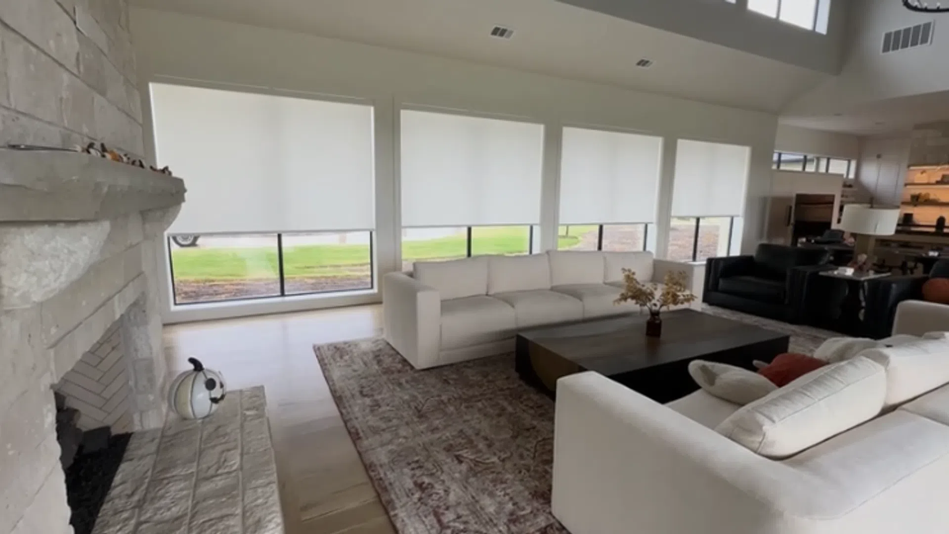 Modern living room with white sofas, large windows with roller blinds, stone fireplace, and wooden coffee table.