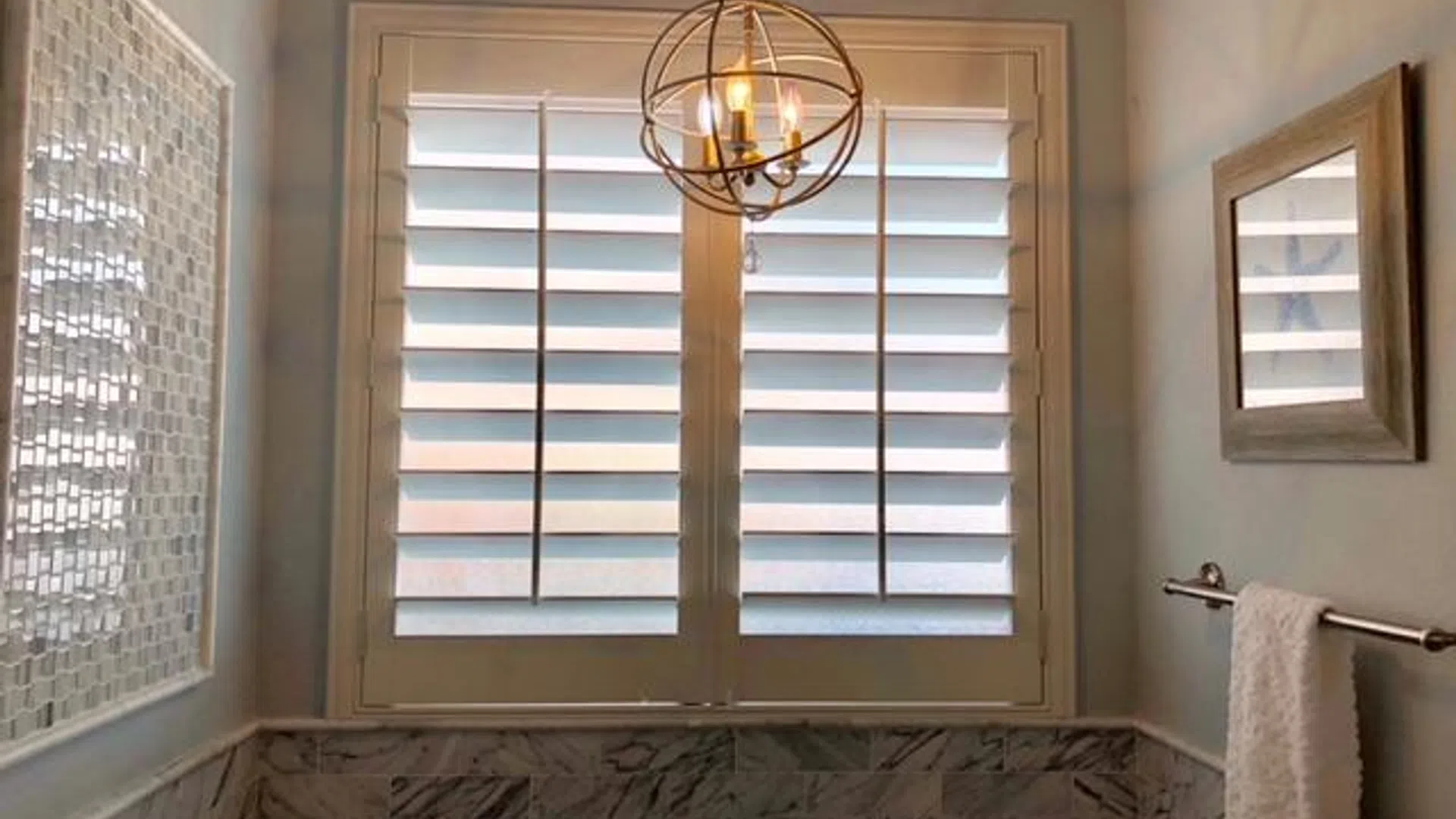 Modern bathroom with marble tiles, white shutters on window, hanging chandelier, towel rack, and framed wall art.