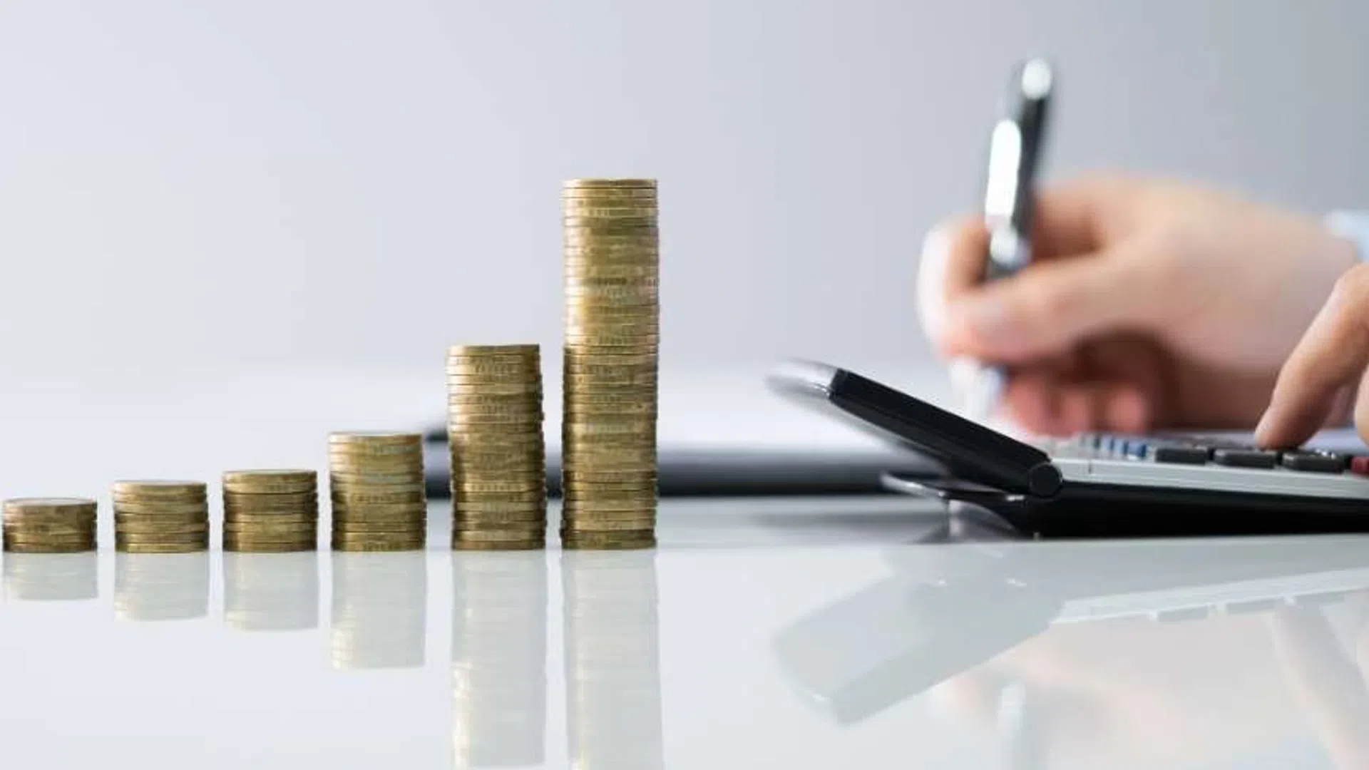 Stacked coins arranged in ascending order next to a person using a calculator and pen for financial calculations.