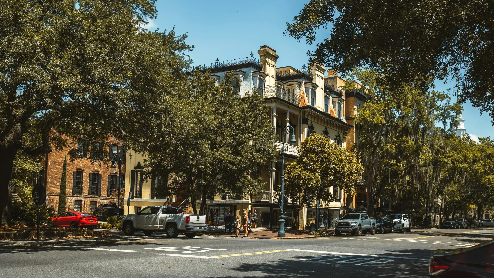 Historic urban street with vintage buildings, large trees, parked cars, and bus stop under clear blue sky.