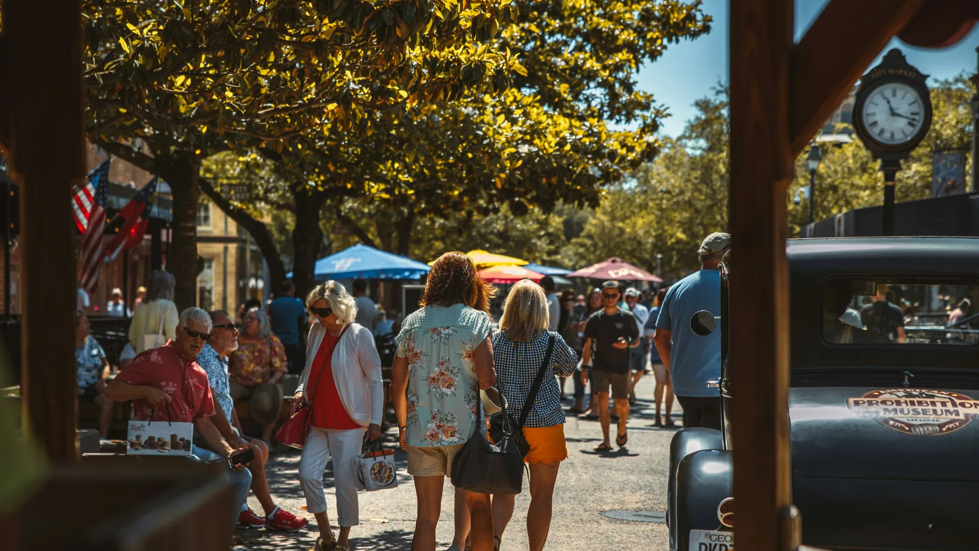 People walking and sitting at an outdoor street market with leafy trees and colorful umbrellas on a sunny day.
