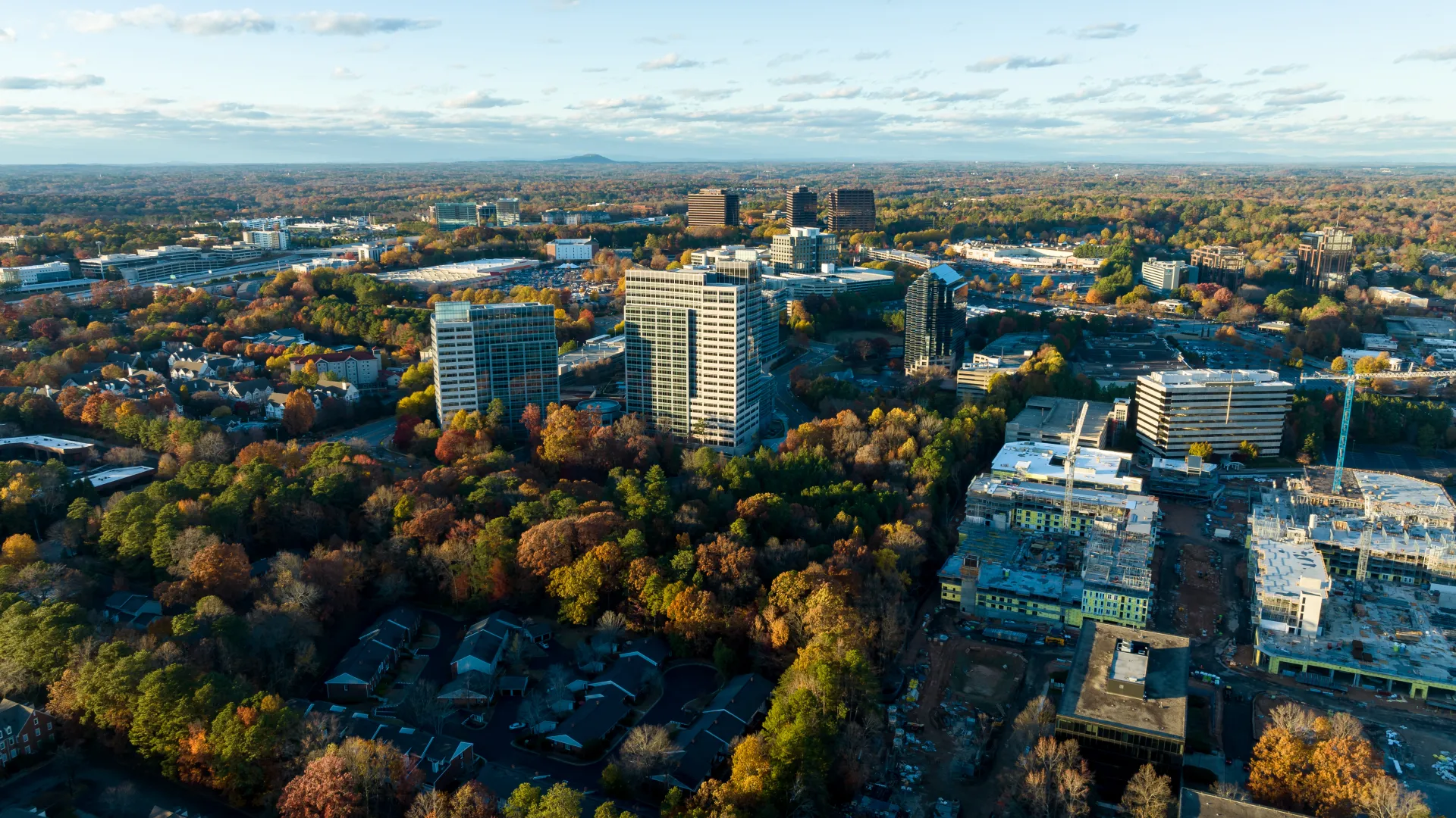 Aerial view of a cityscape with modern buildings surrounded by trees in autumn under a partly cloudy sky.