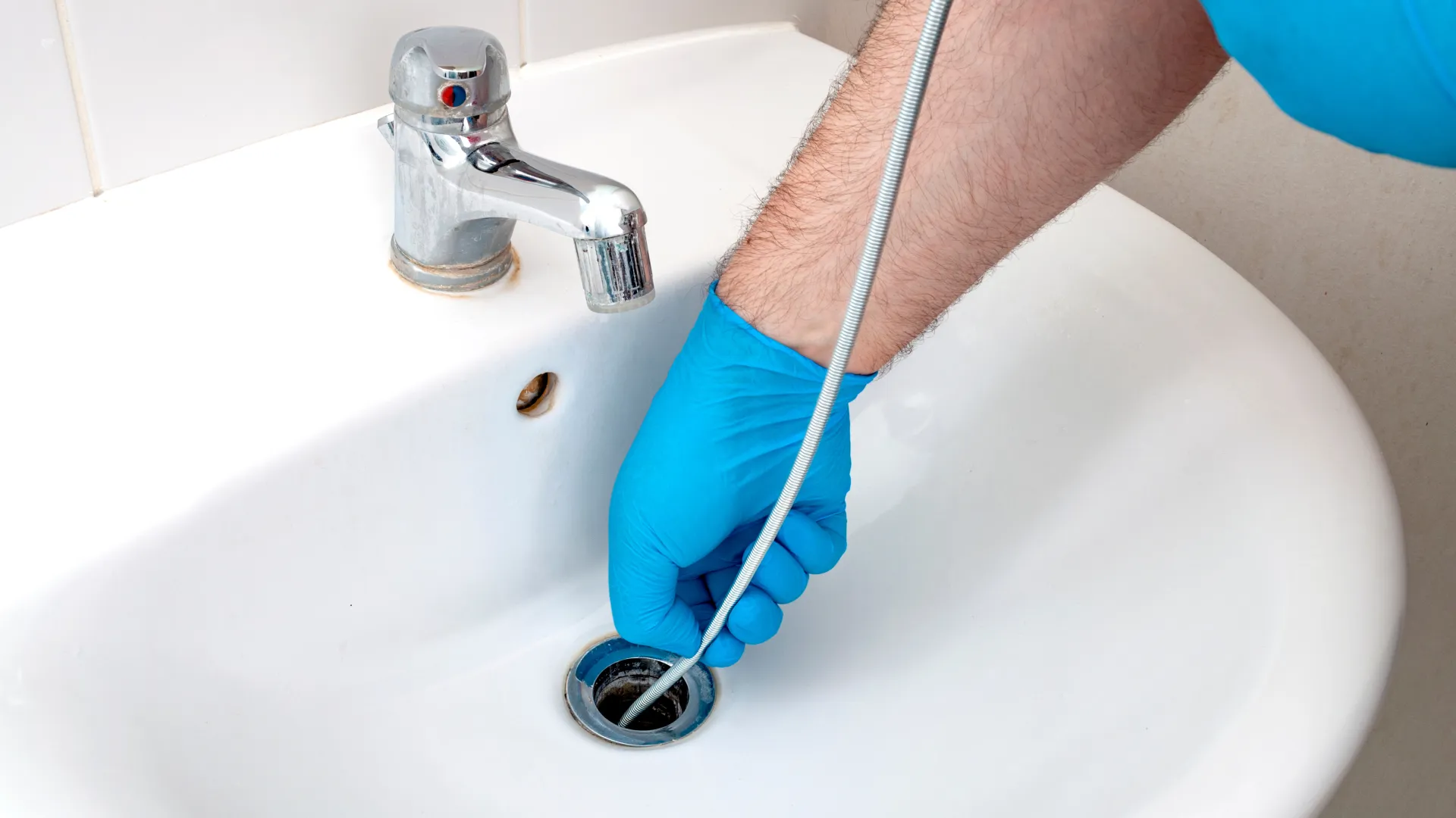 Person wearing blue gloves unclogs a white sink drain using a plumbing snake tool.