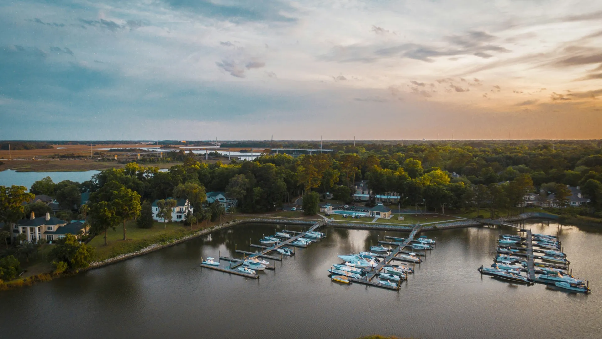 Aerial view of a marina with boats docked along piers surrounded by trees and houses at sunset.