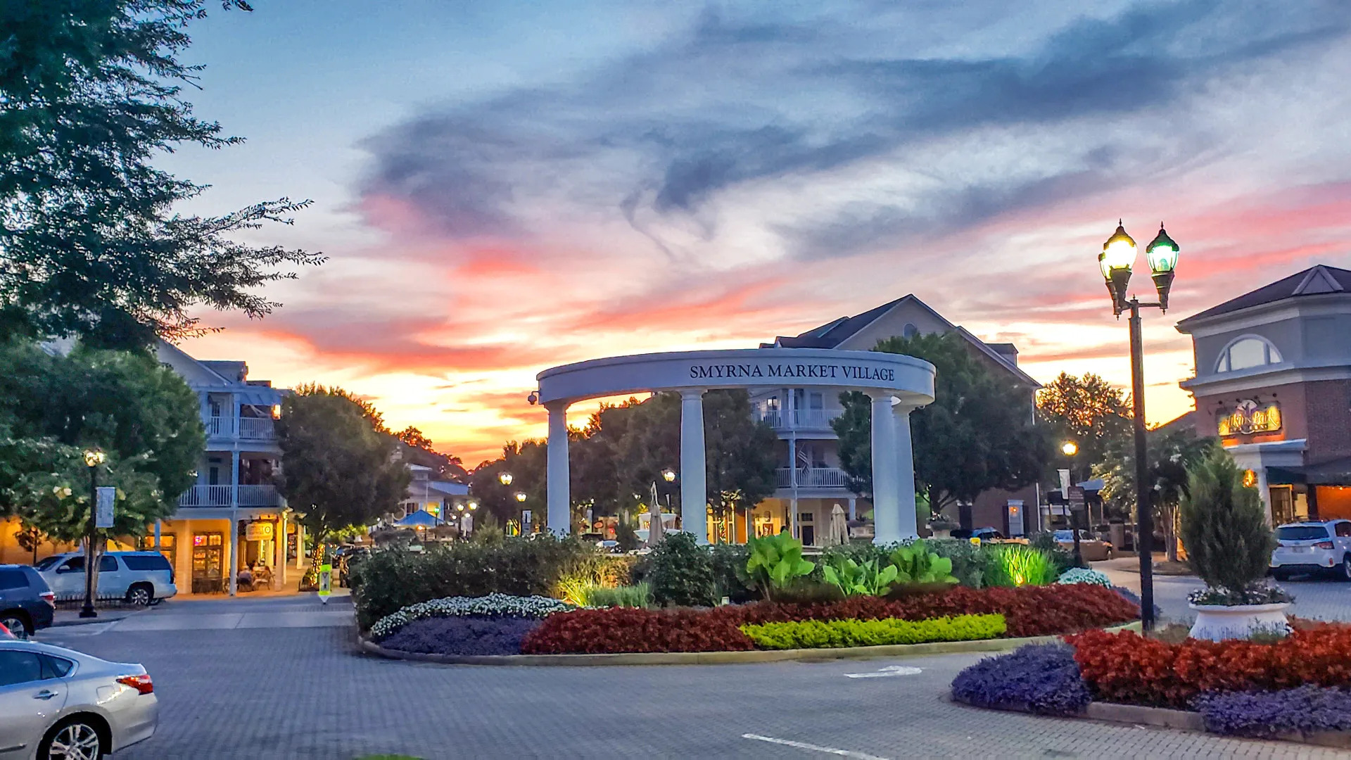 Smyrna Market Village entrance at sunset with colorful sky, landscaped roundabout, and street lamps glowing.
