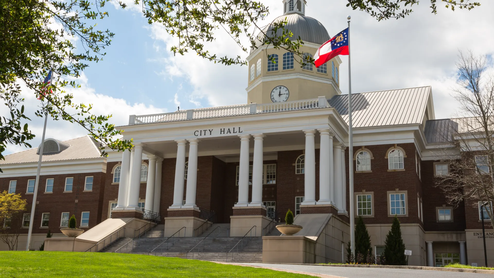 City hall building with classical columns, clock tower, and Georgia state flag against a blue sky.