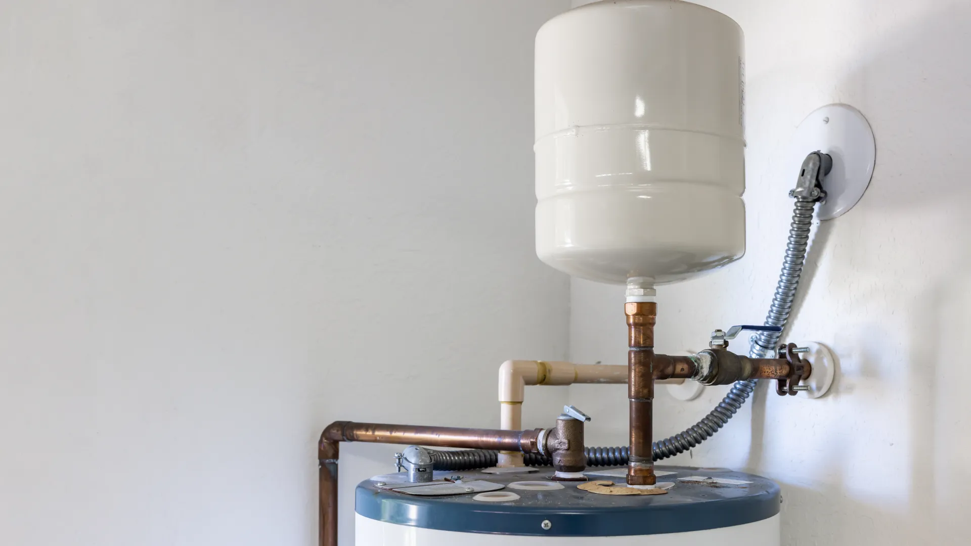 Home water heater tank with expansion tank and copper piping installed on white wall indoors.