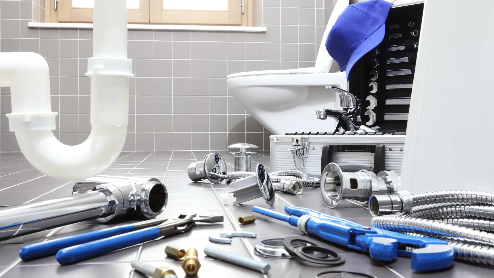 Plumbing tools and parts laid out on tiled bathroom floor with toilet and toolbox in background