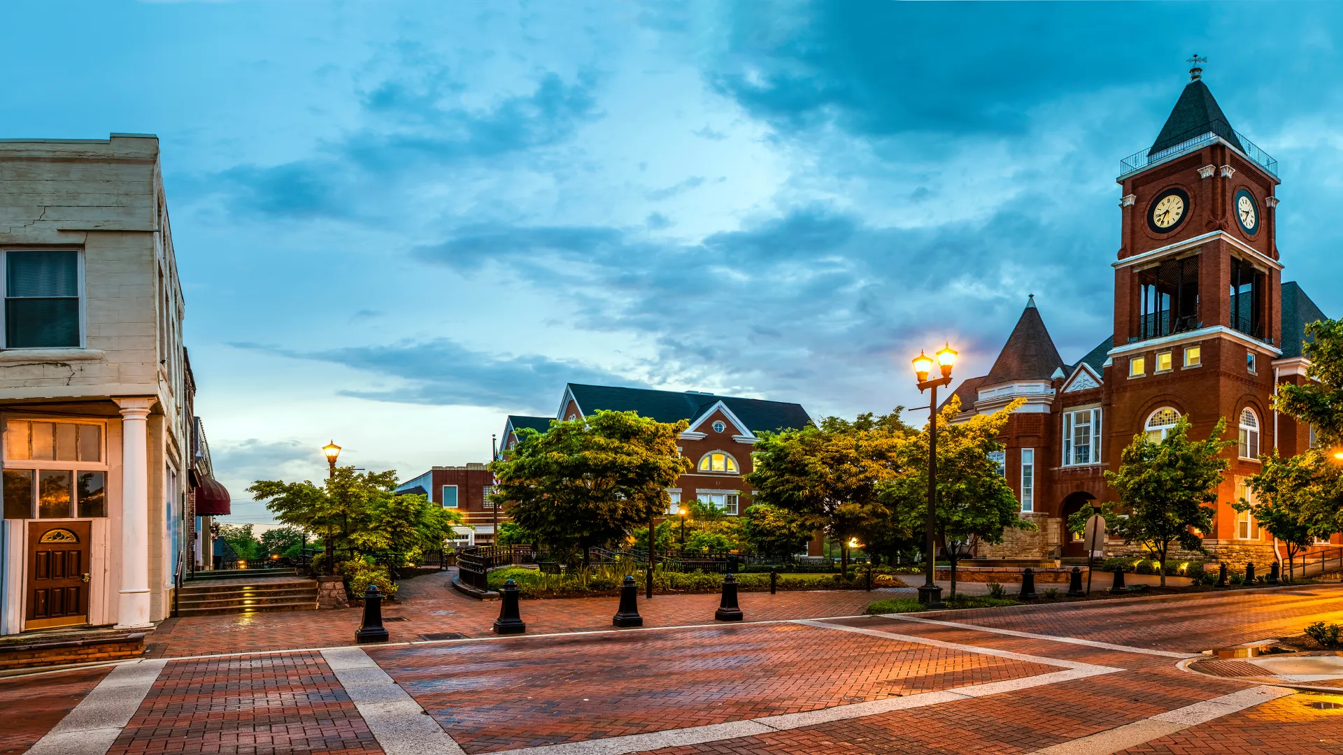 Historic town square with brick sidewalks, lampposts, trees, and a clock tower at dusk under blue sky.