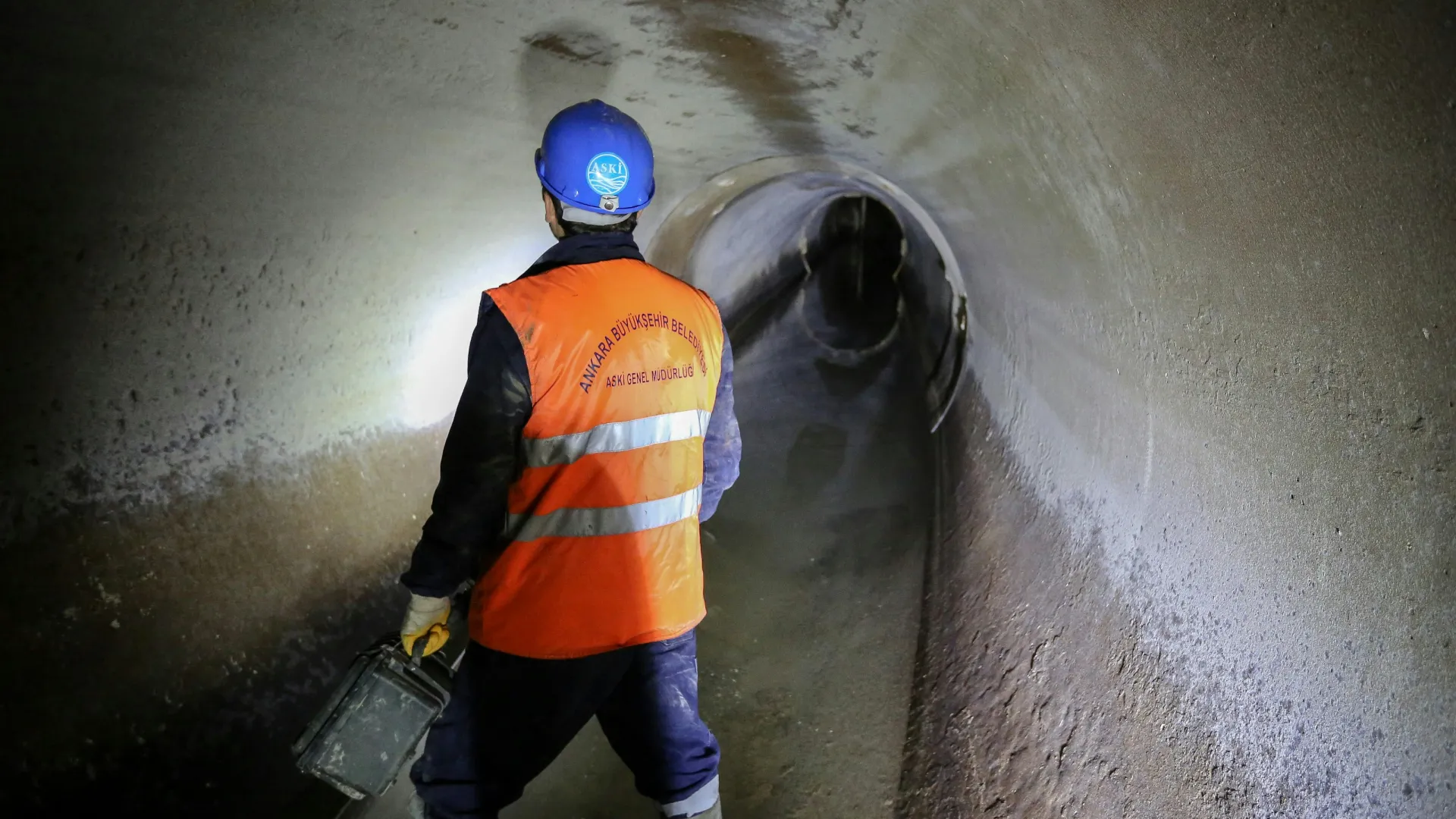 Worker in orange vest and blue helmet inspecting a tunnel with water on the floor, using a flashlight.