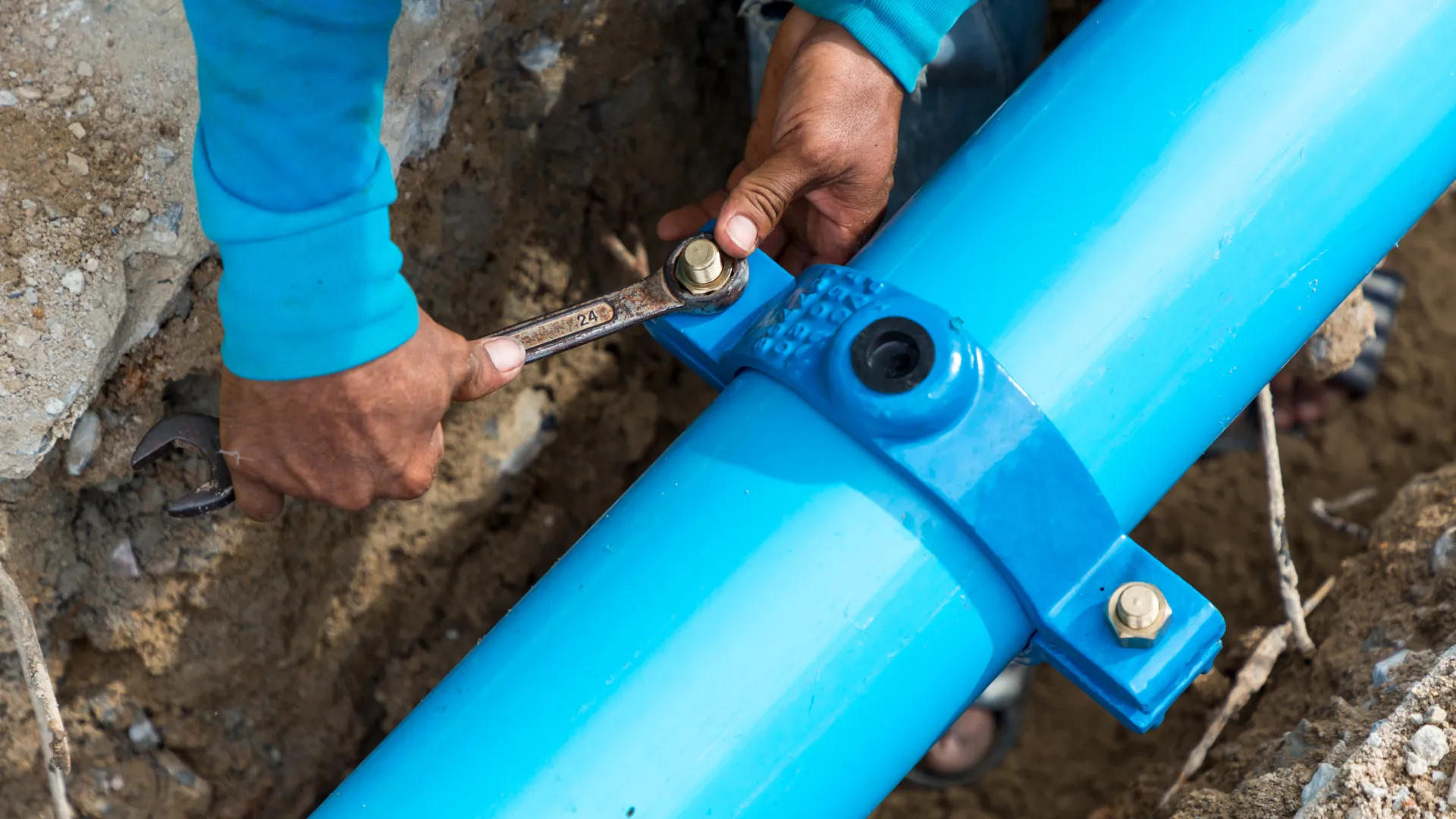 Worker tightening bolt on large blue underground water pipe using wrench in construction site trench