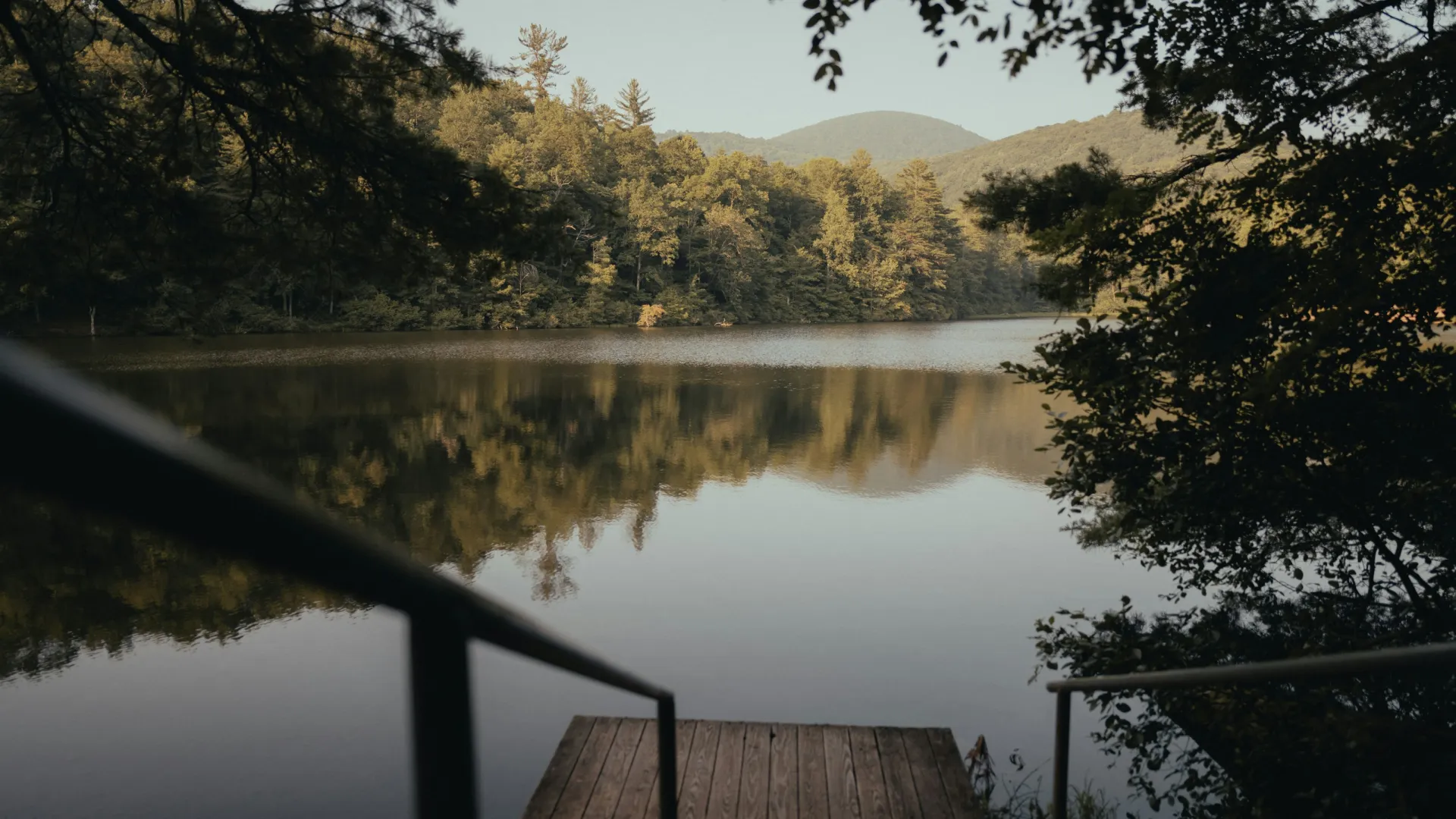 Wooden dock with metal railings overlooking calm lake reflecting forested mountains and trees at sunset.