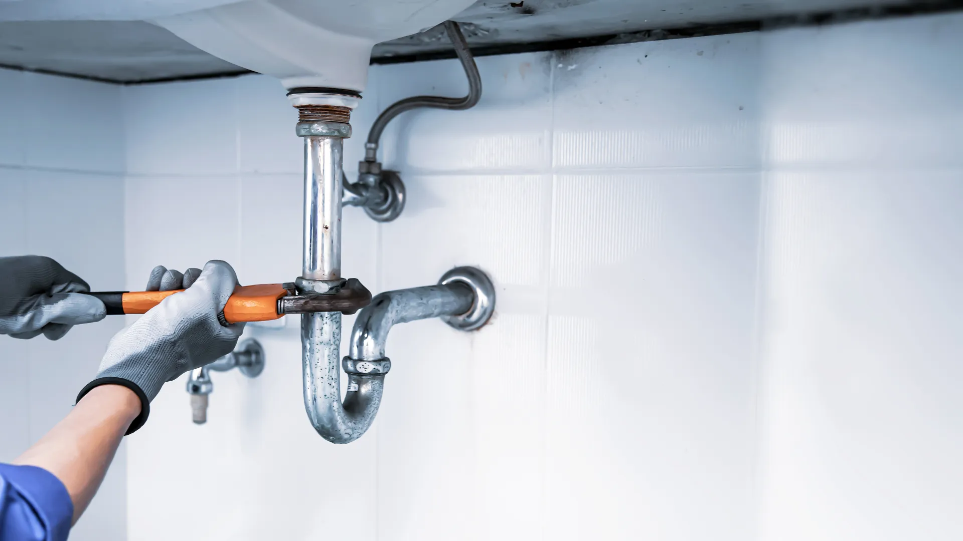 Plumber wearing gloves fixing a pipe under a sink with a wrench in a tiled bathroom setting.
