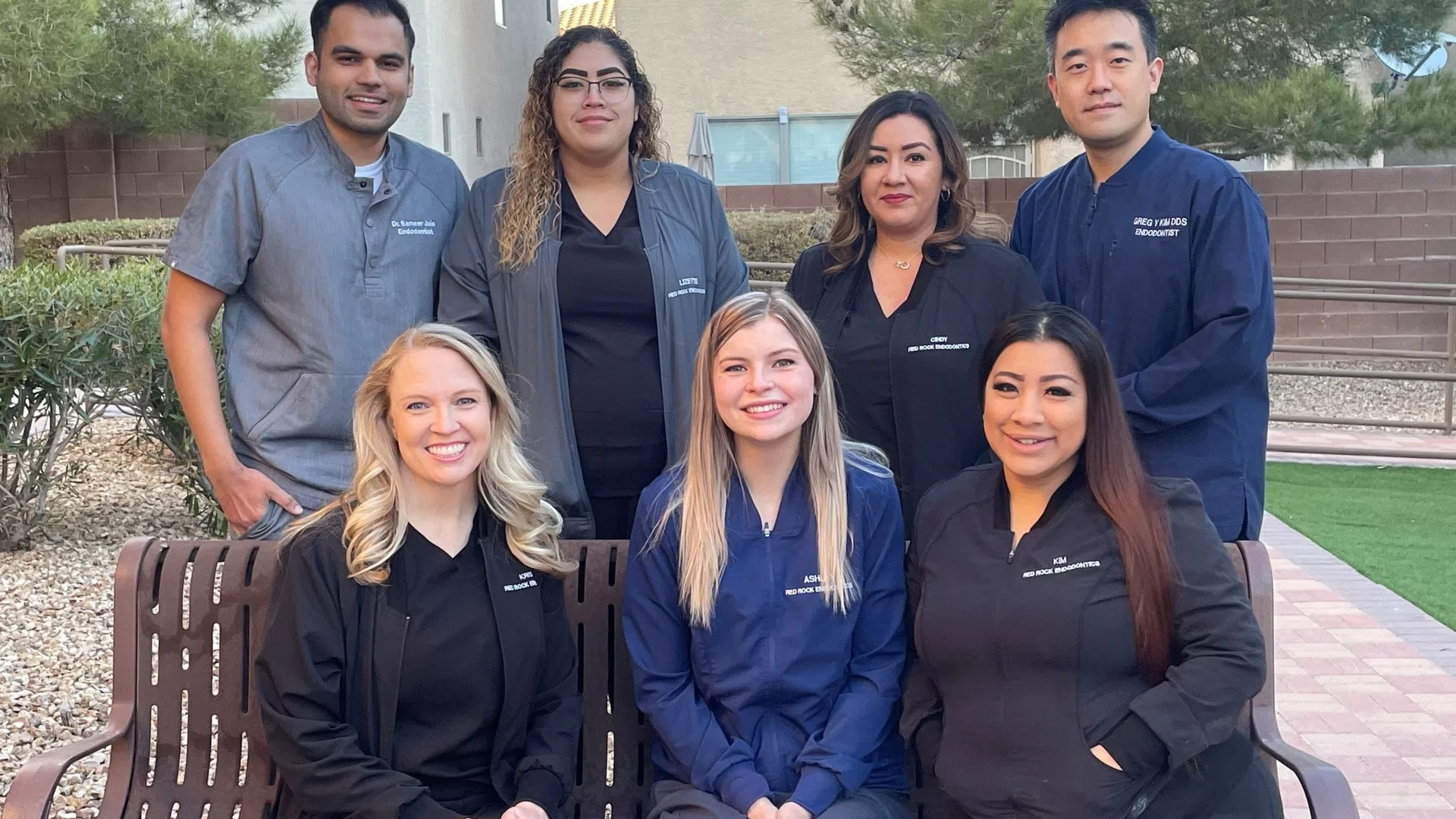 Group of seven diverse dental professionals smiling outdoors in uniforms sitting and standing near bench.
