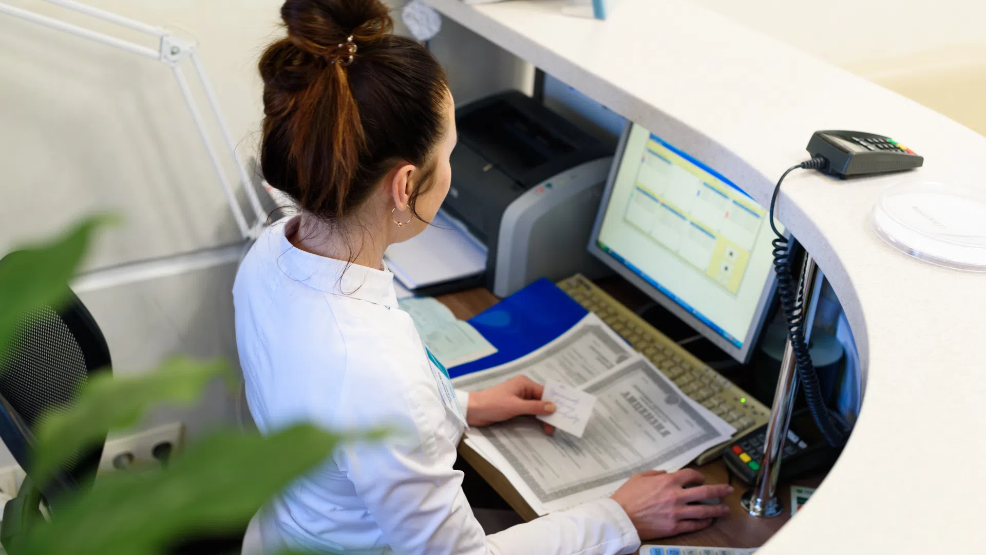 Woman in white coat working at a reception desk with computer, documents, and card reader.