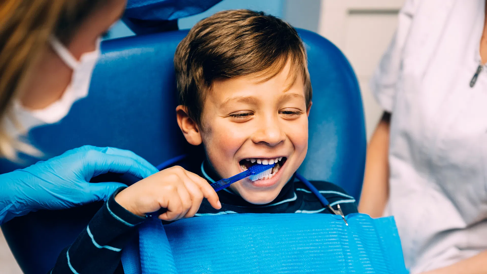 Young boy smiling and brushing teeth at dental clinic with dentist assisting, wearing blue gloves and dental bib.