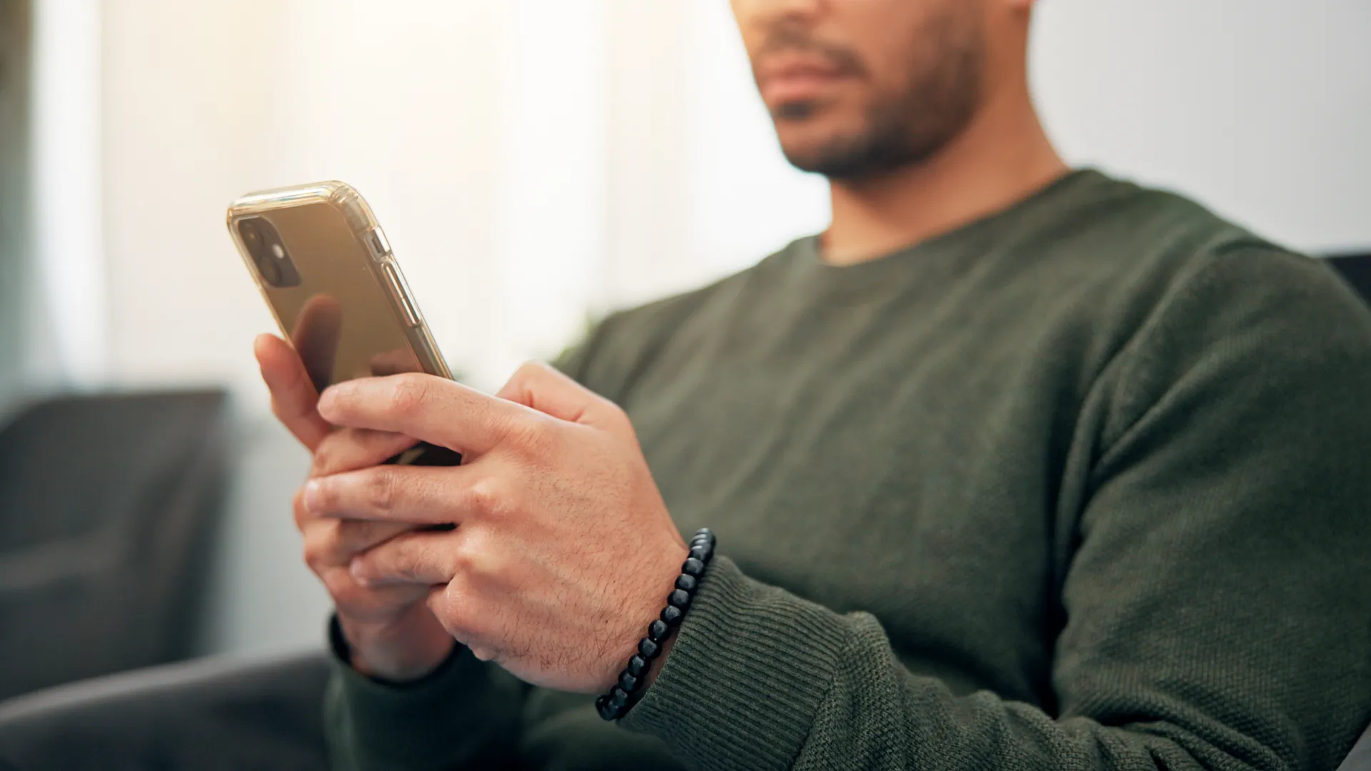 Man wearing dark green sweater using smartphone indoors with bracelet on wrist, blurred background.