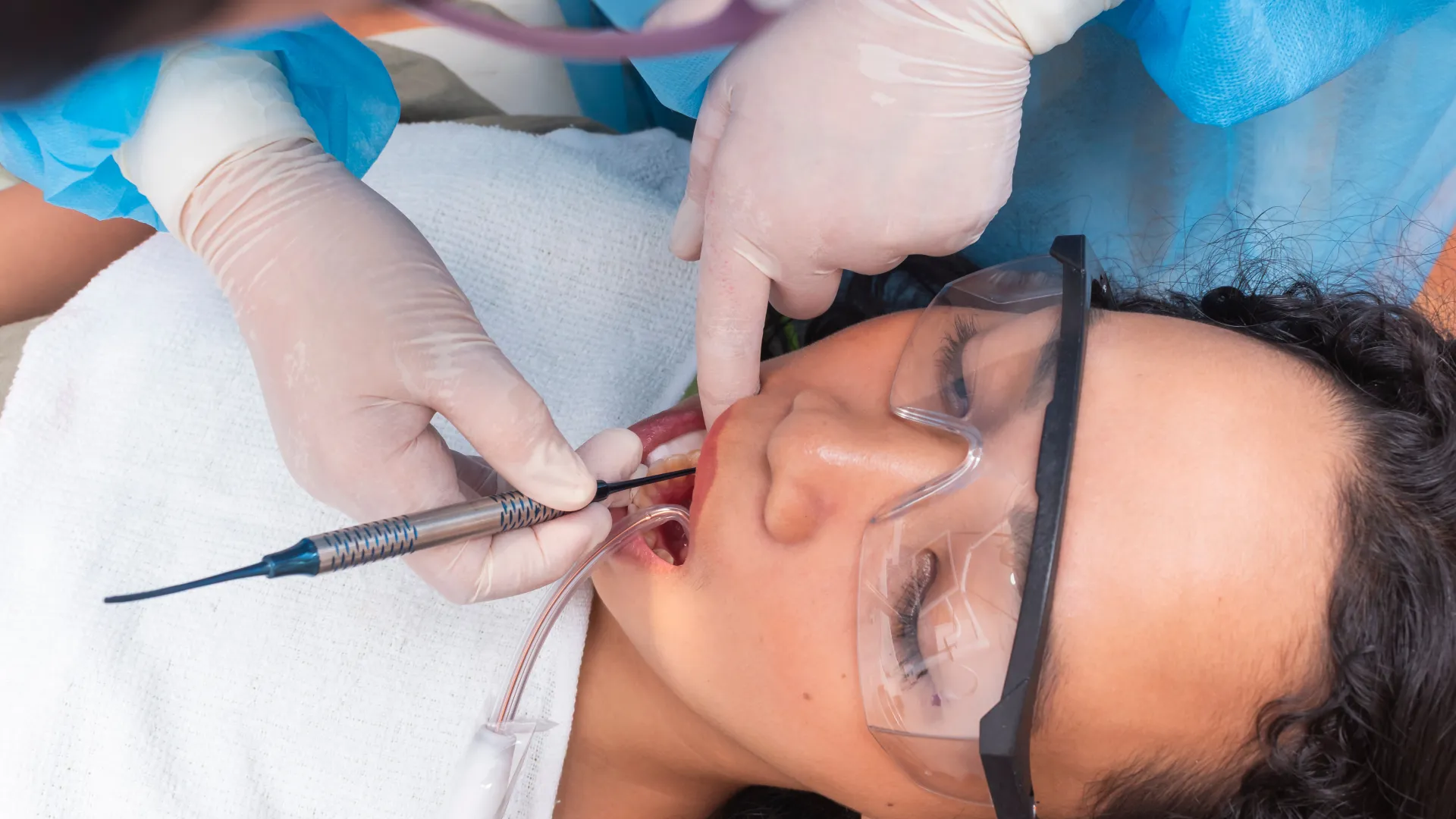 Dentist wearing gloves treating patient's teeth using dental tools while patient wears protective glasses.