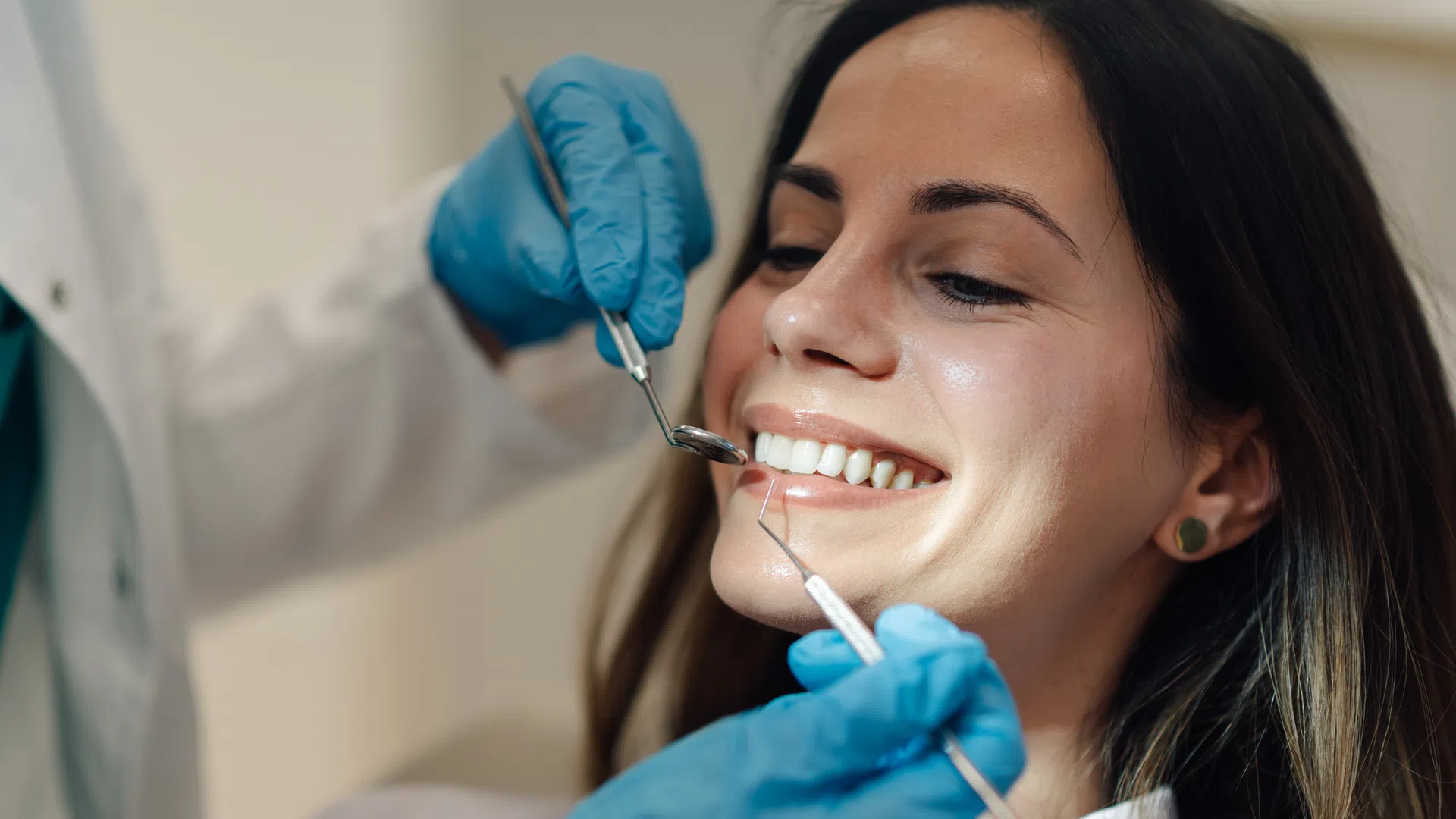 Patient smiling while dentist wearing blue gloves examines teeth with dental tools during checkup