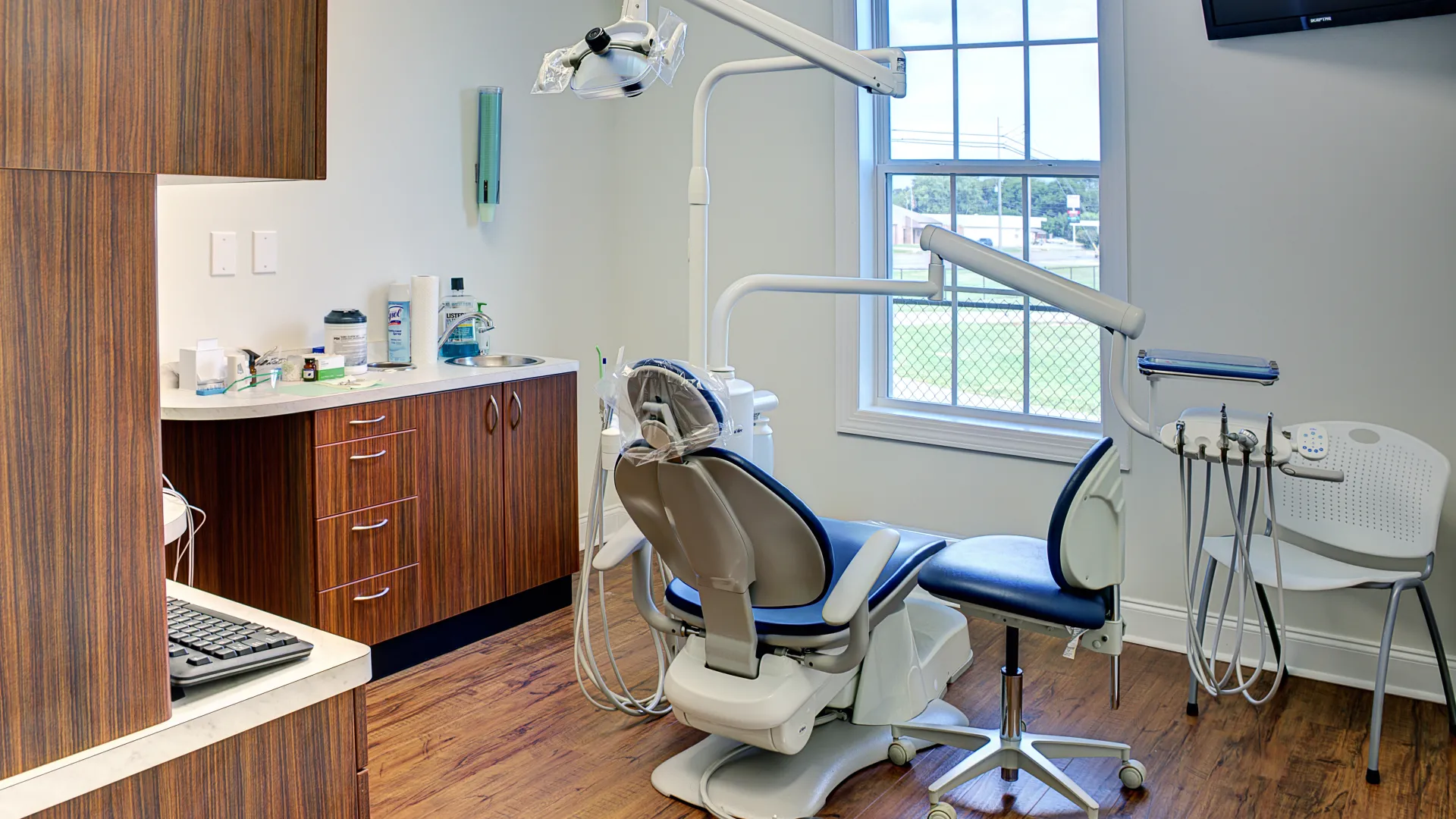 Modern dental clinic room with chair, dental equipment, wooden cabinets, and a window showing an outdoor view.