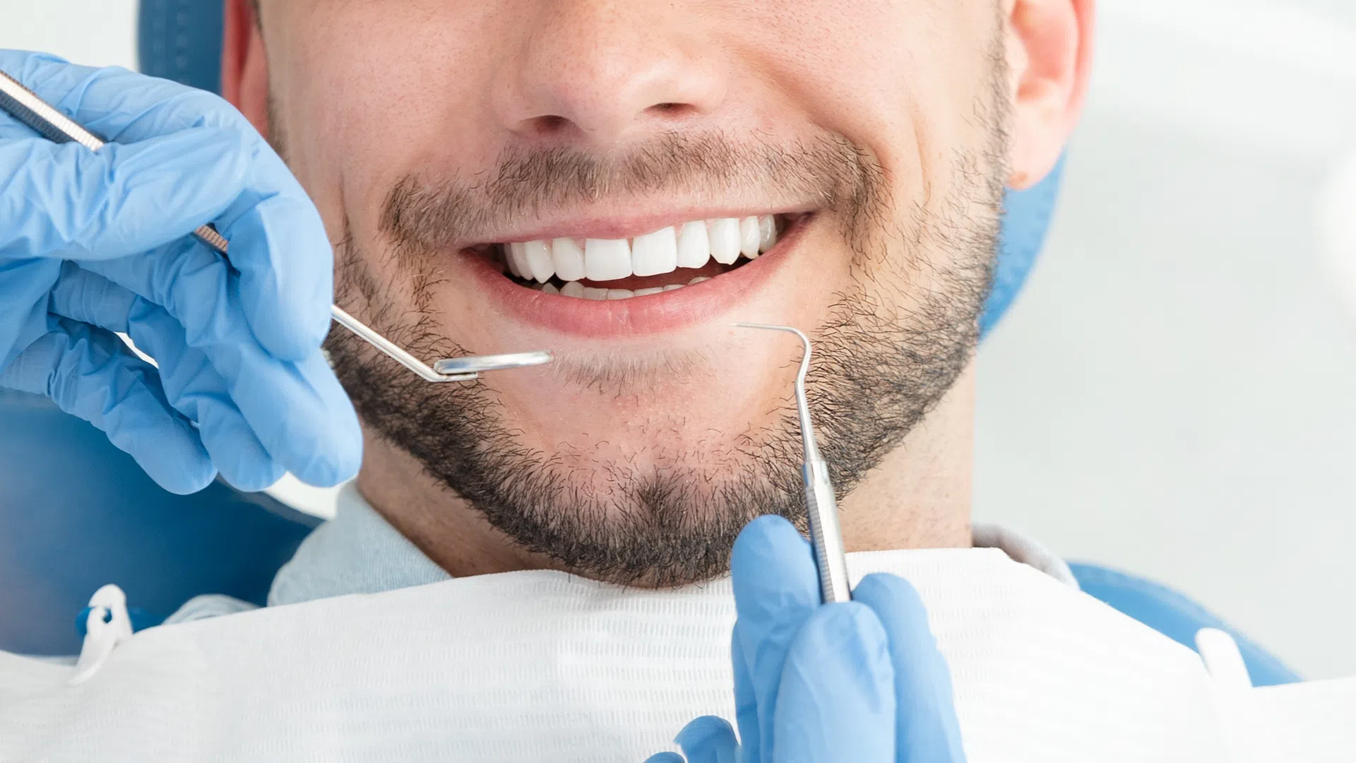 Dentist wearing blue gloves examining a smiling man's teeth with dental tools in a clinic.