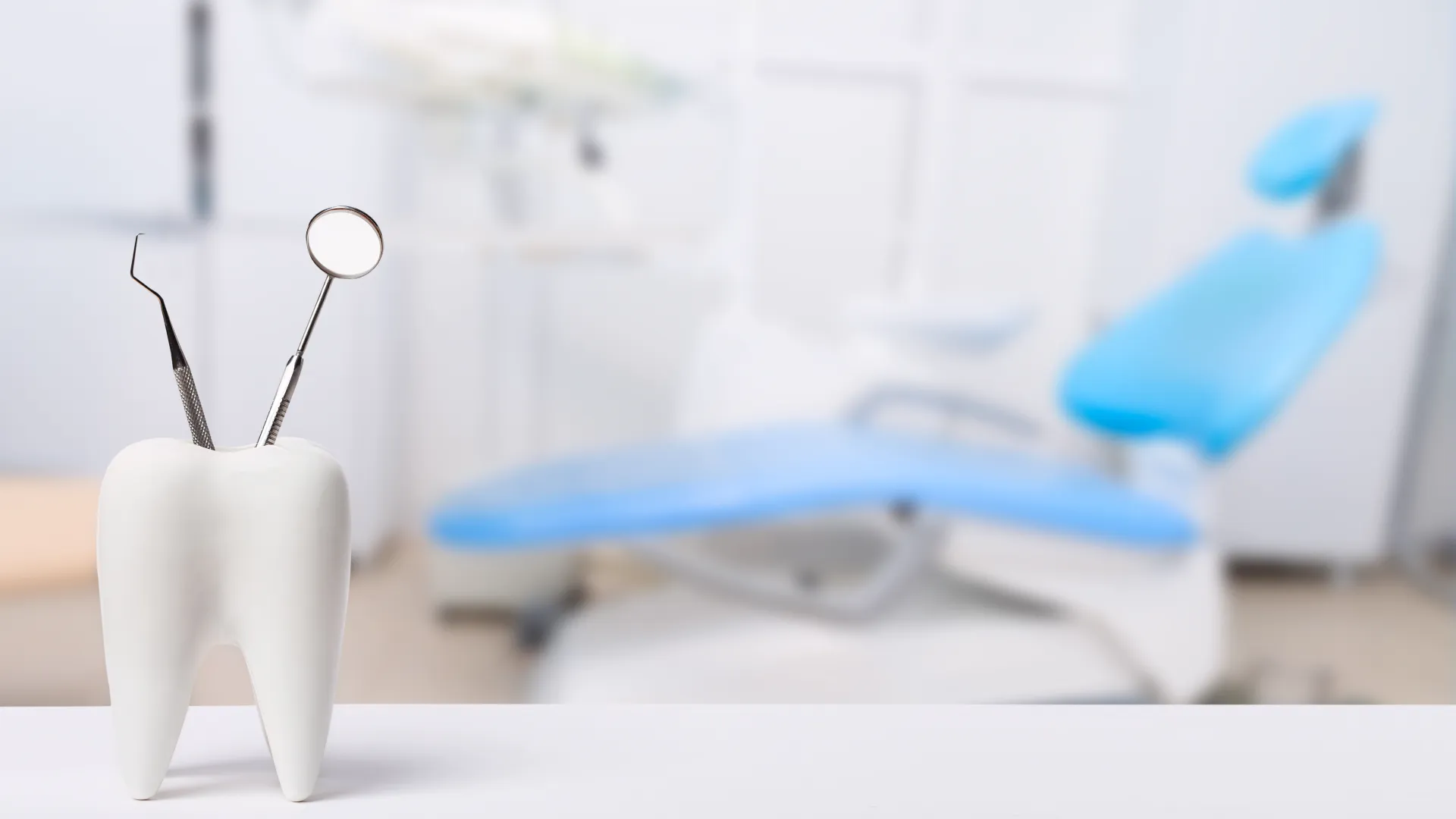Dental tools in a tooth-shaped holder with a blue dental chair in the blurred background of a clinic.