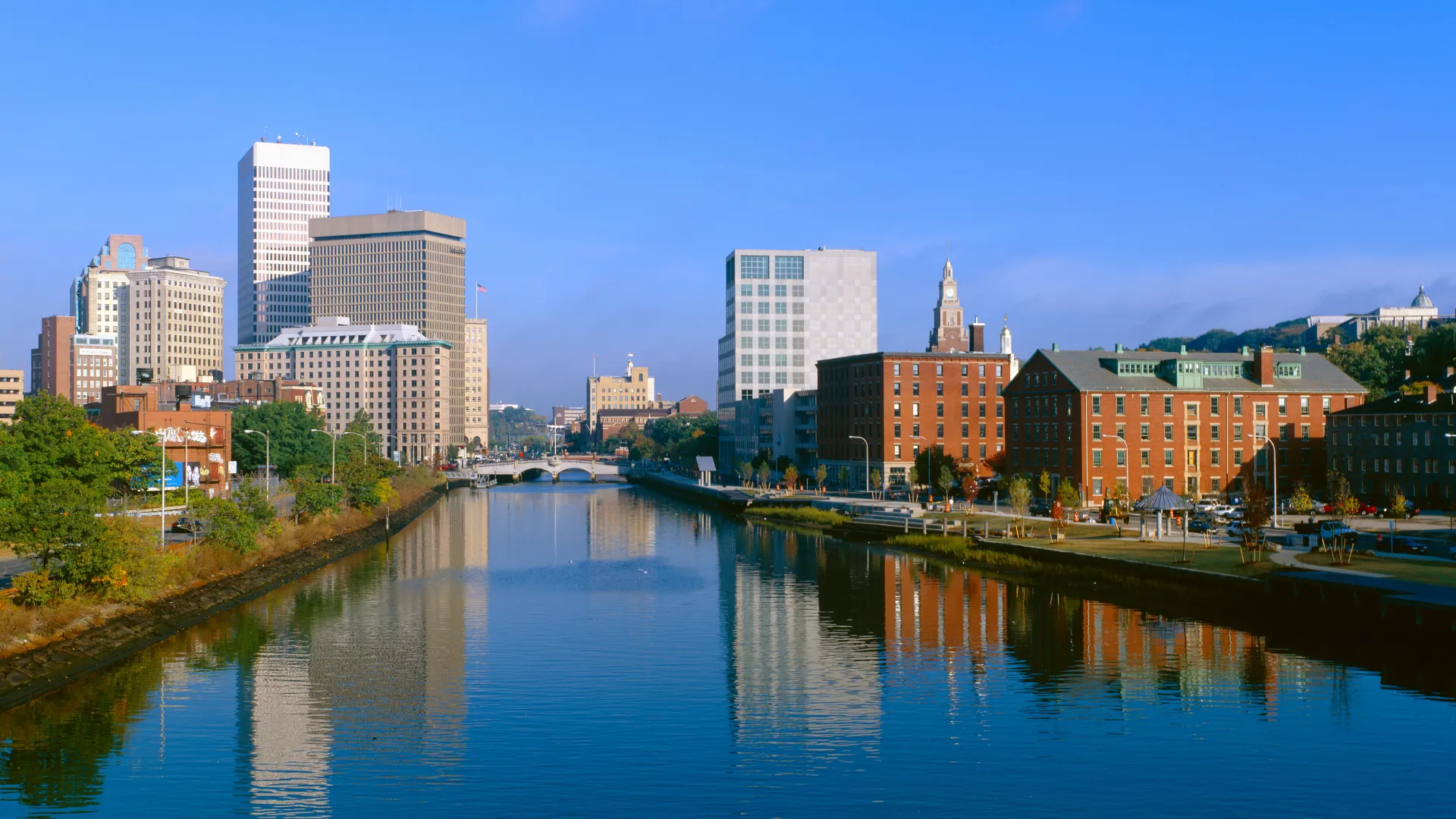 City skyline with diverse buildings reflected in a calm river under a clear blue sky.
