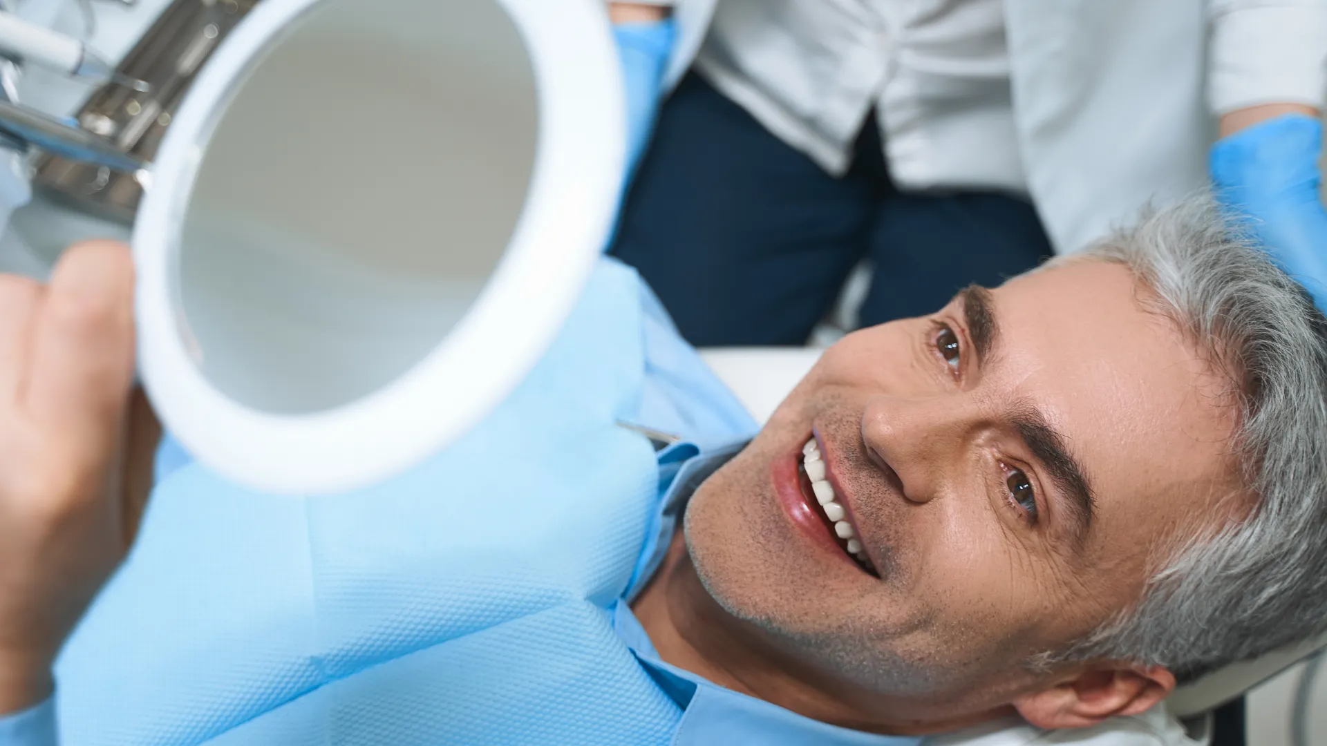 Middle-aged man smiling at his reflection in a mirror during a dental checkup with a dentist.