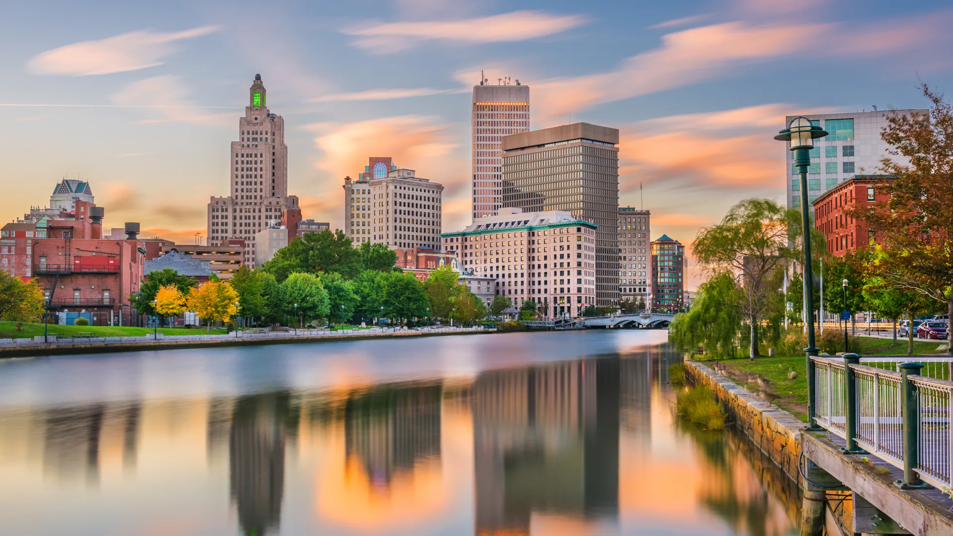 City skyline with tall buildings reflecting on calm river during colorful sunset with trees and riverside path.