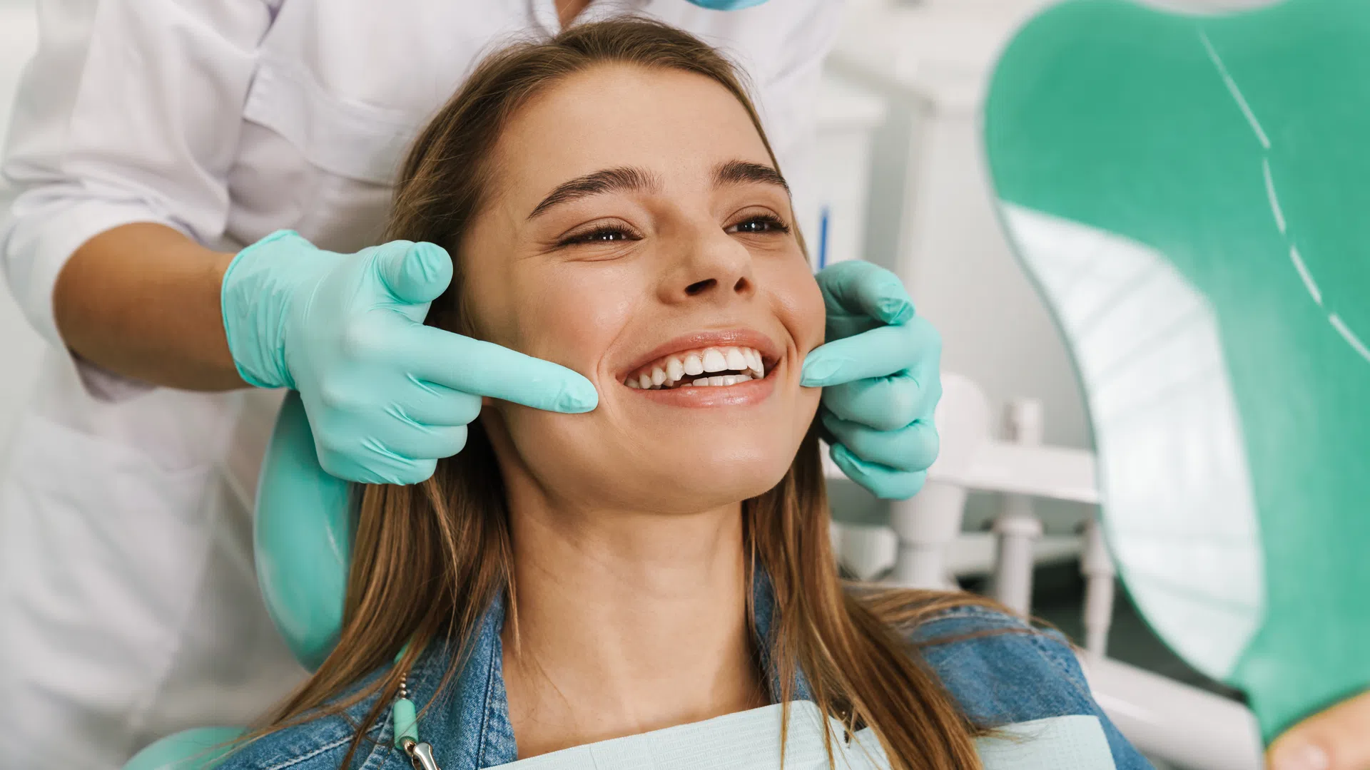 Dentist wearing gloves examines a smiling woman's teeth using a mirror in a dental clinic.