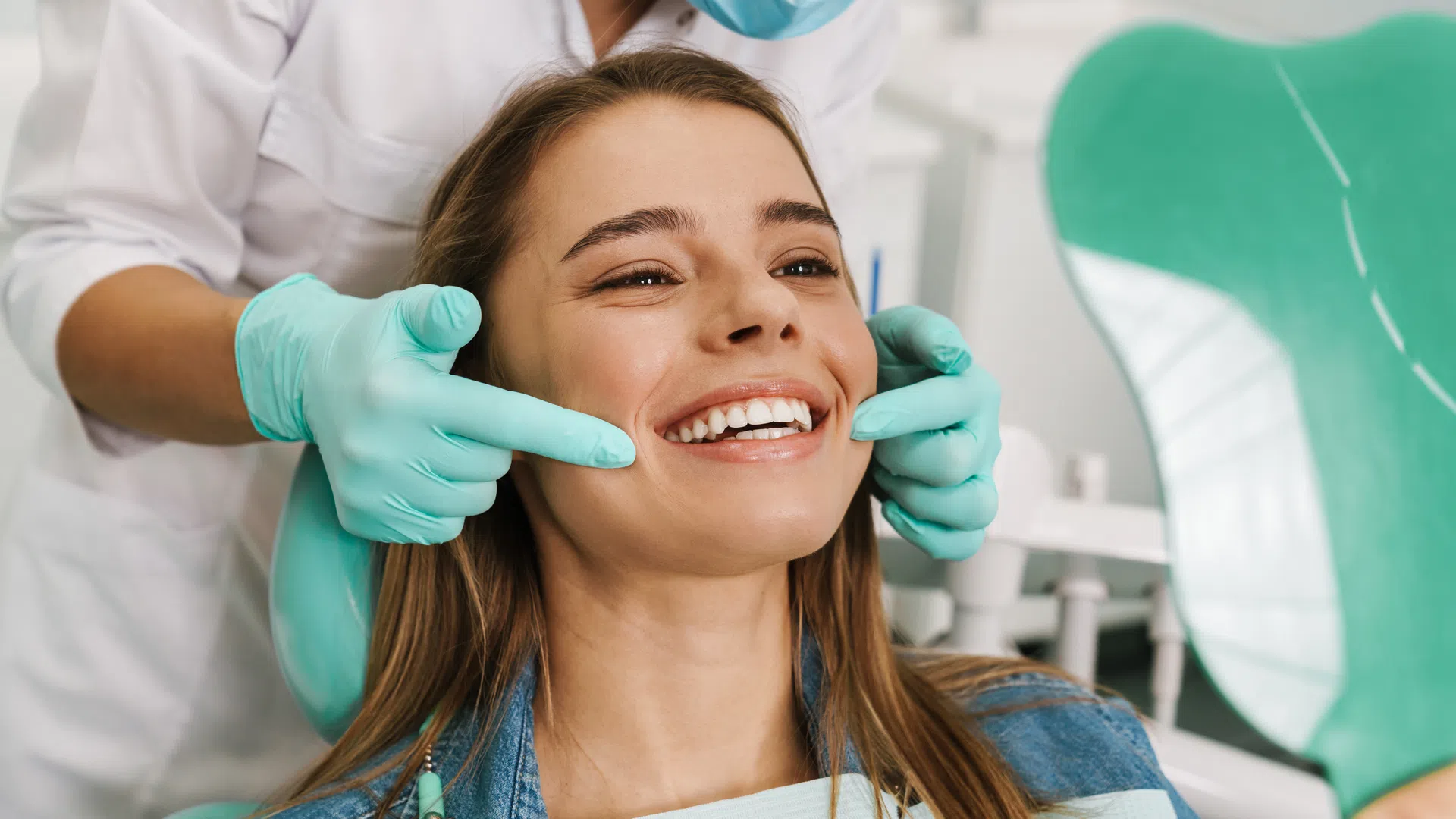 Dentist wearing gloves examines a smiling woman's teeth using a mirror in a dental clinic.