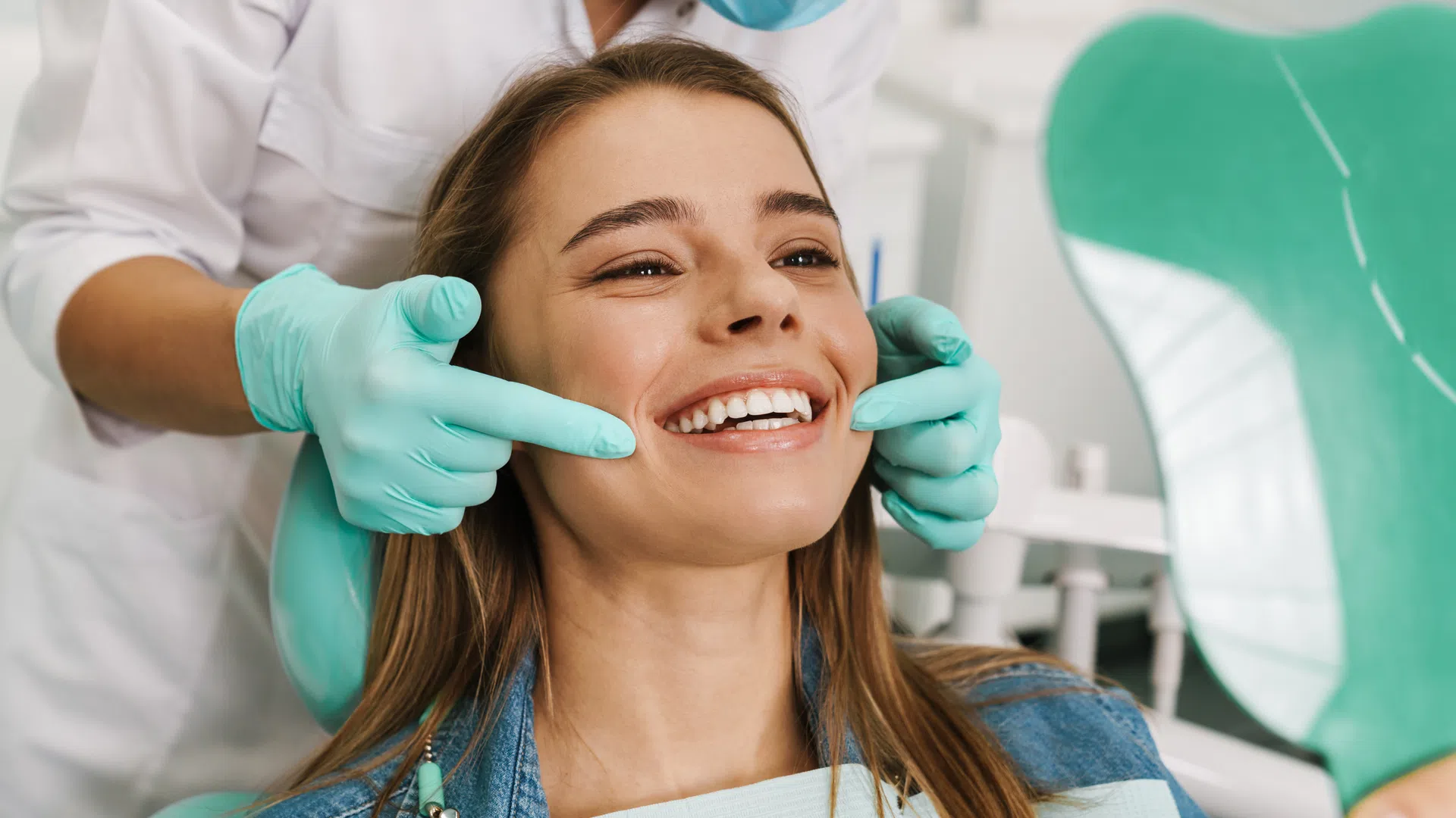 Dentist wearing gloves examines a smiling woman's teeth using a mirror in a dental clinic.