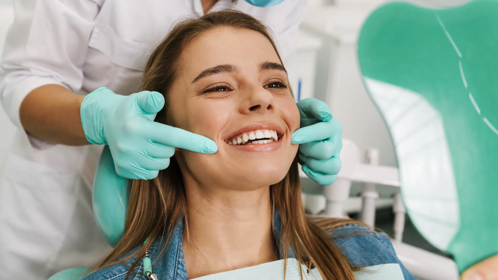 Dentist wearing gloves examines a smiling woman's teeth using a mirror in a dental clinic.