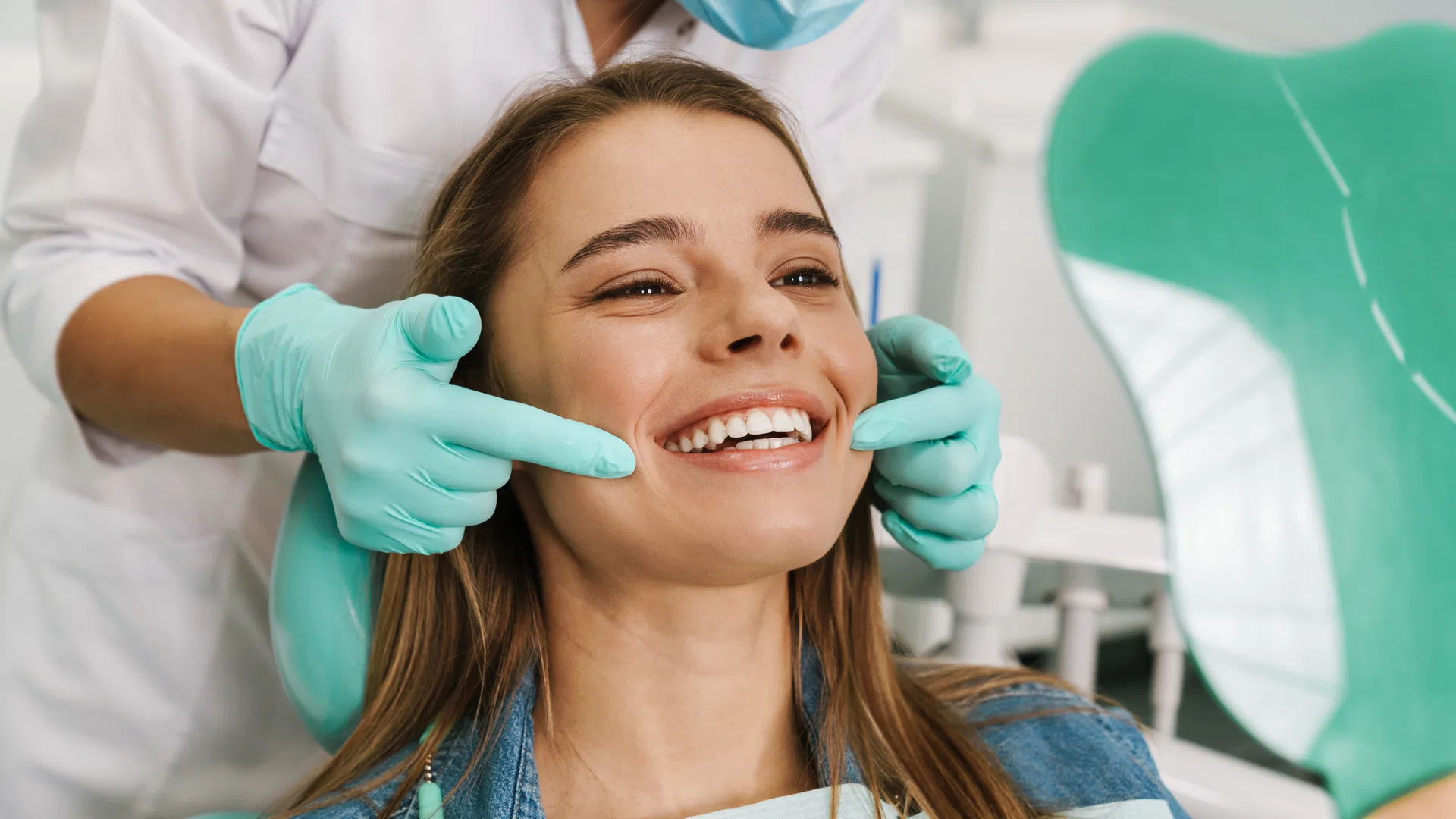 Dentist wearing gloves examines a smiling woman's teeth using a mirror in a dental clinic.