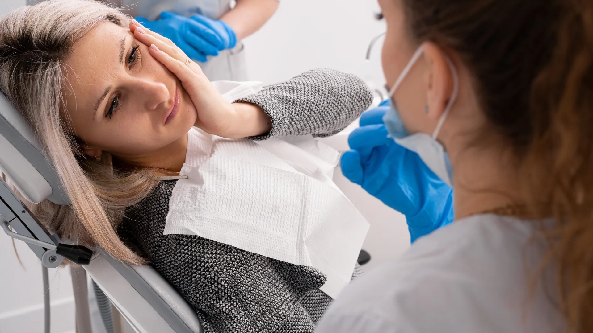 Female patient in dental chair holding her cheek in pain while dentist wearing blue gloves examines her.