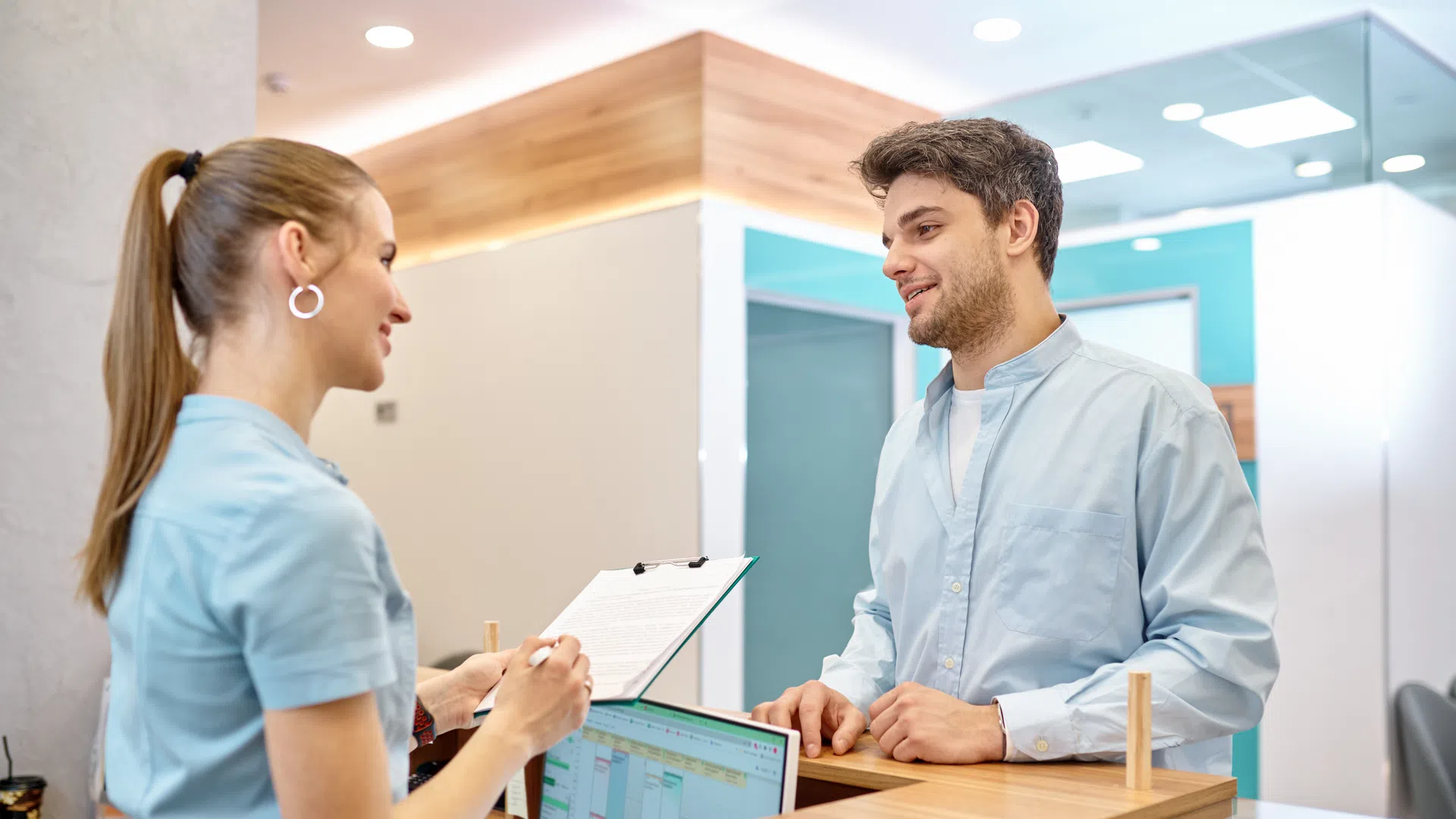 Young man talking to smiling receptionist with clipboard at medical clinic reception desk
